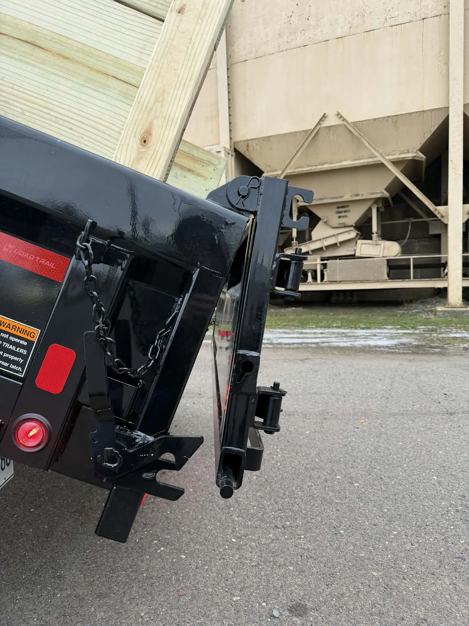 Close-up of the hitch and safety chains on a black trailer with a load of wooden planks, with a large industrial hopper or silo in the background.