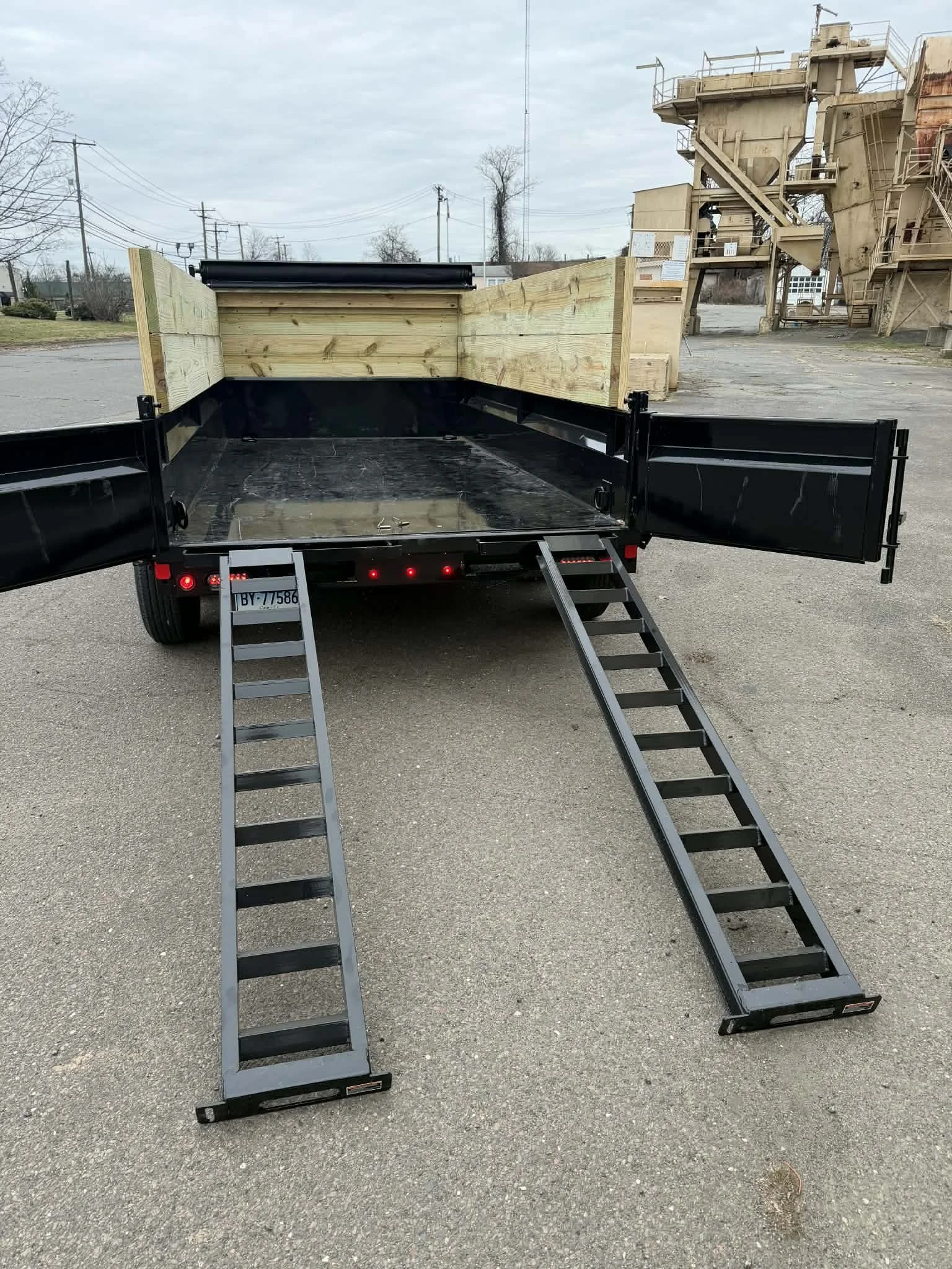 A black flatbed trailer with two foldable ramps, showing the open rear with side walls and a wooden back wall, parked on a paved lot with an industrial building in the background.