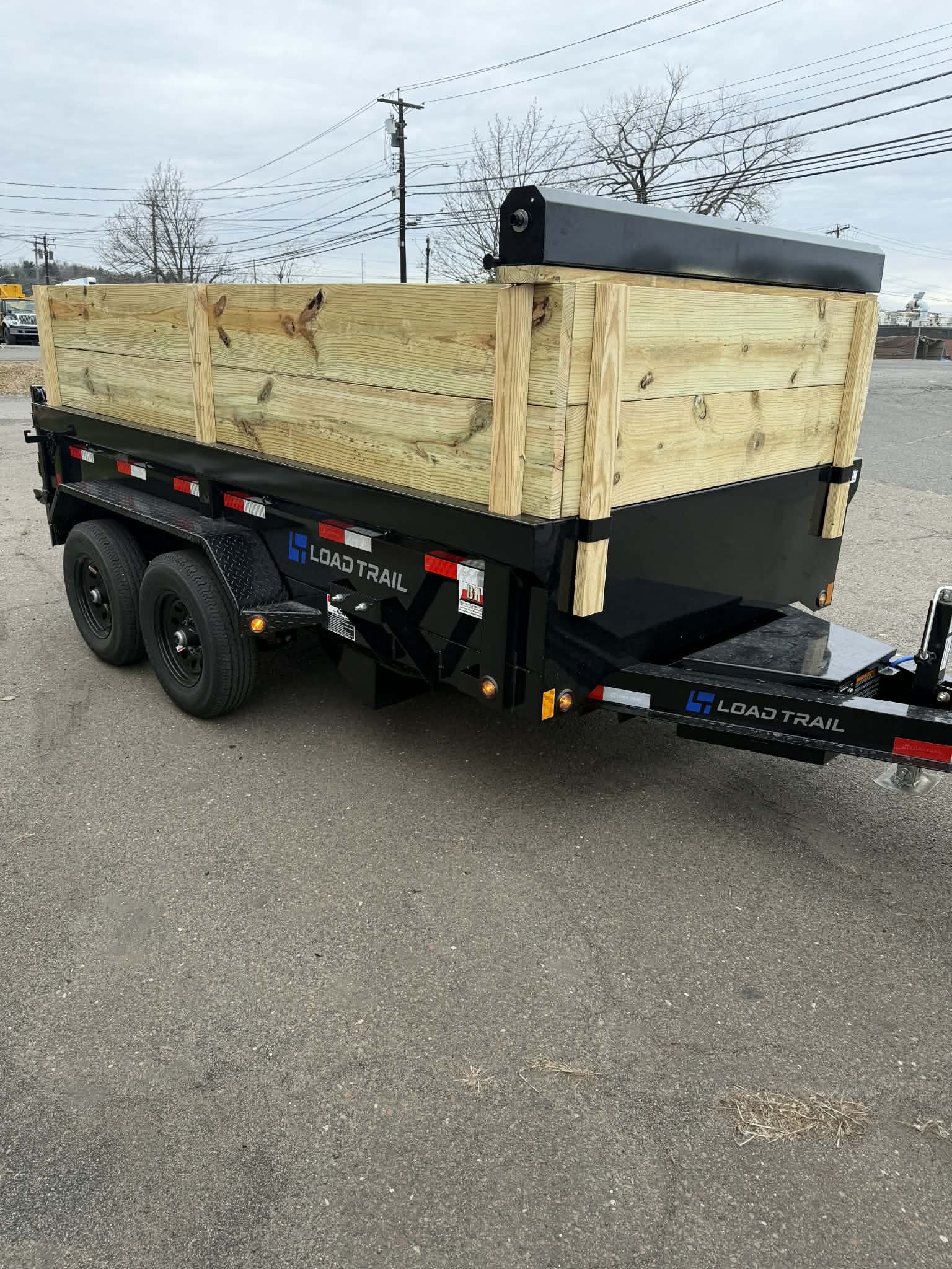 A Load Trail utility trailer carrying a large wooden crate in an outdoor lot with overhead power lines and trees in the background.