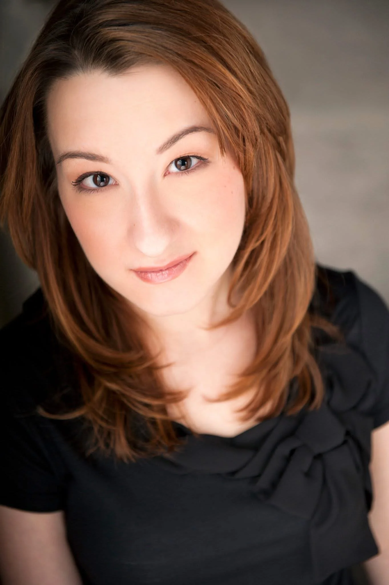 Close-up of a woman with auburn hair, wearing a black top, smiling subtly and looking at the camera.