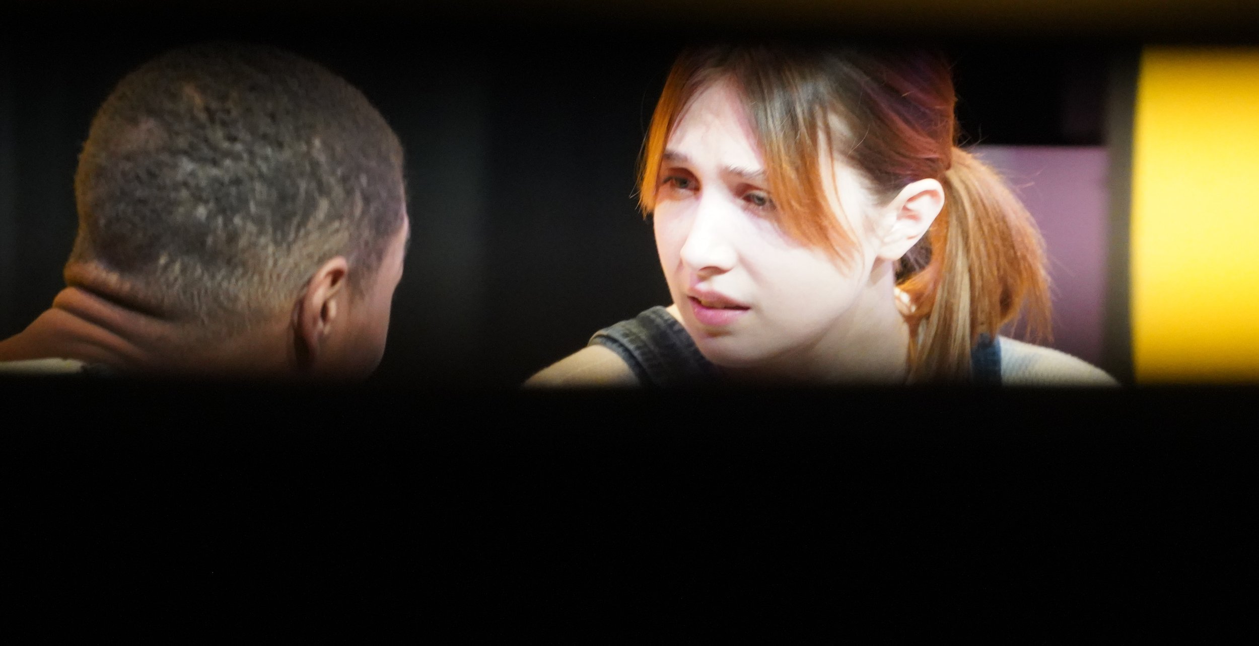 A young woman with red hair talking to a man with short hair, viewed through a small horizontal opening.