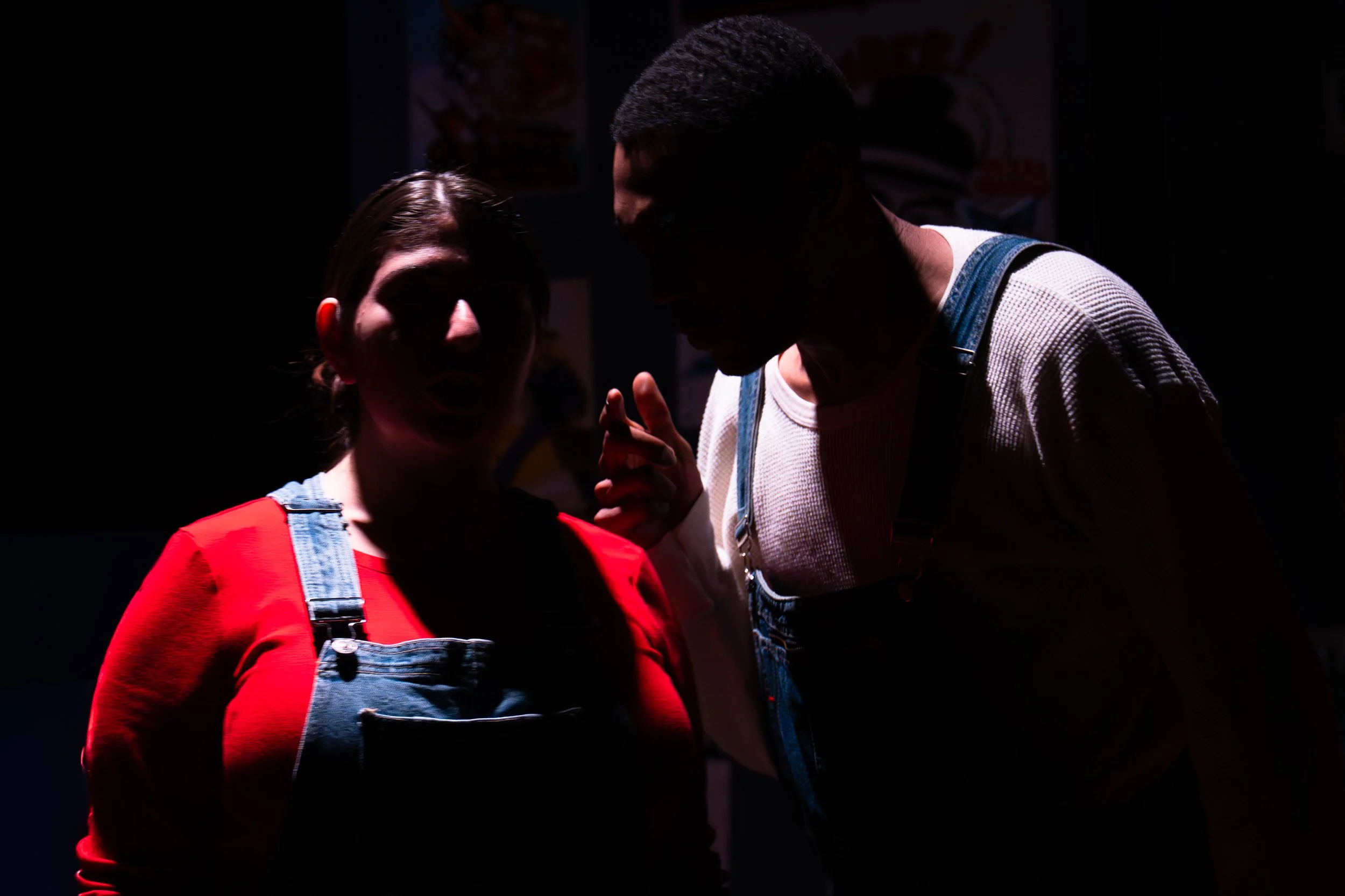 A woman in a red shirt and denim overalls is talking to a man in a white shirt and denim overalls, both in a dark room.