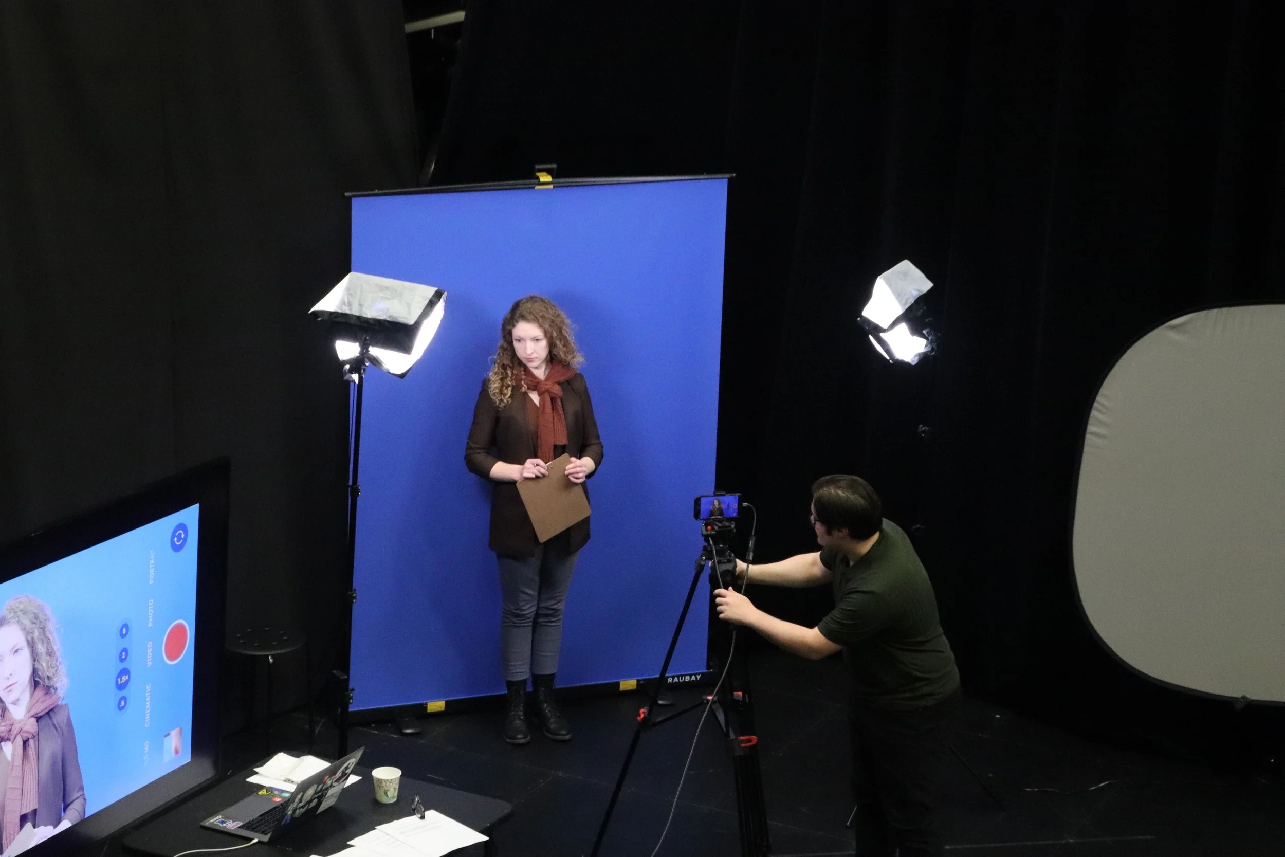 A woman with curly hair standing in front of a blue screen, holding a clipboard, while two men set up photography or video equipment in a studio with professional lighting.