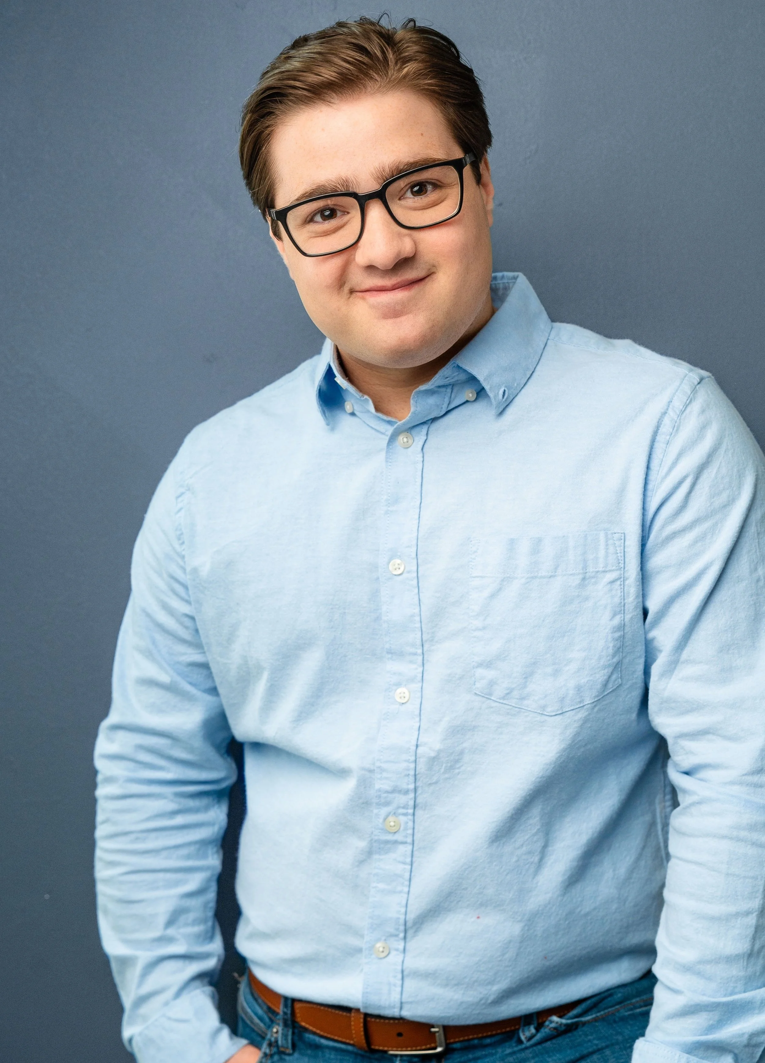 Portrait of a young man with brown hair, wearing black glasses, a light blue button-up shirt, and a brown leather belt, standing against a gray background.