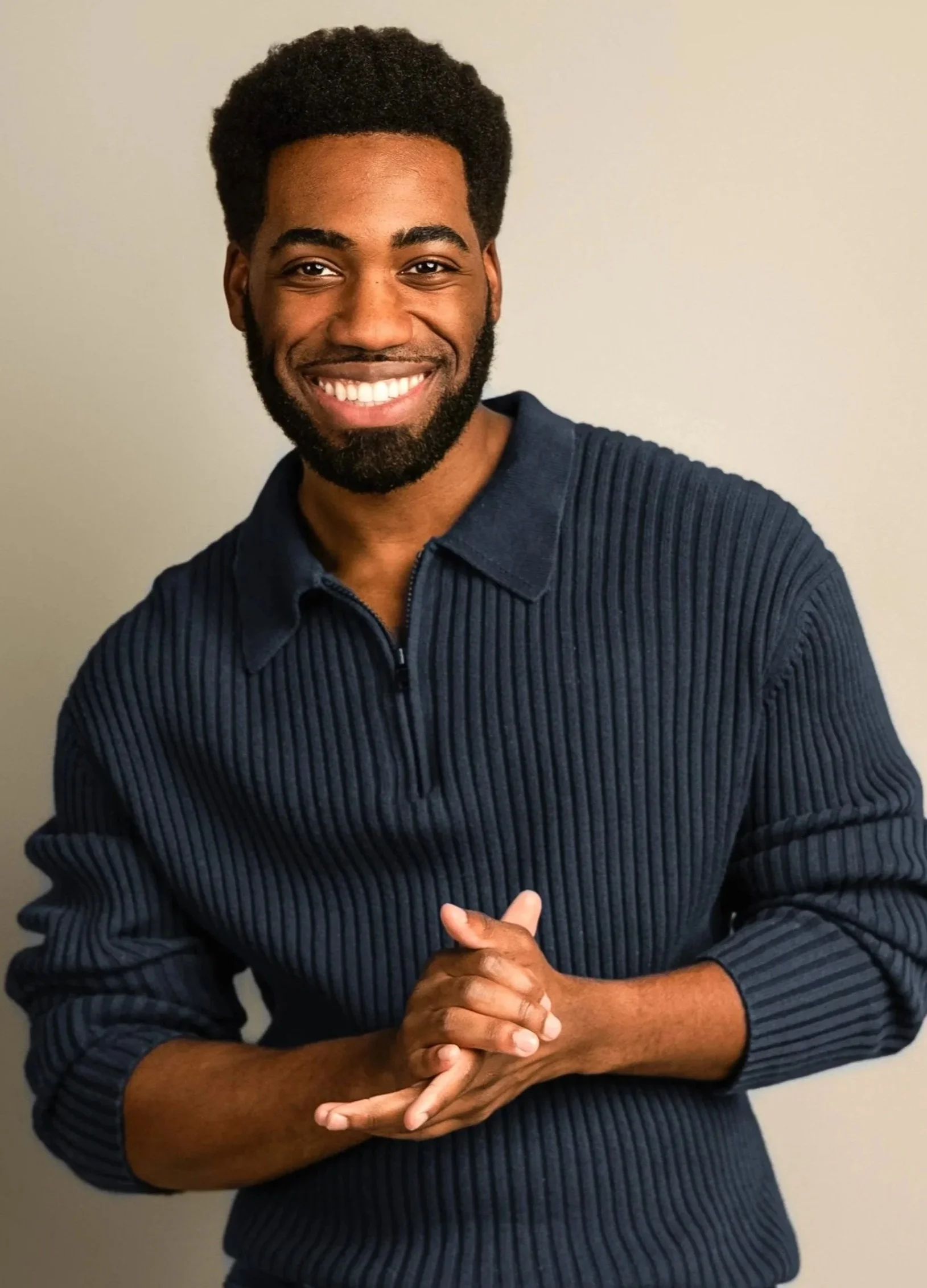 A smiling man with a beard and natural hair, wearing a dark blue ribbed shirt, standing in front of a plain light-colored wall.
