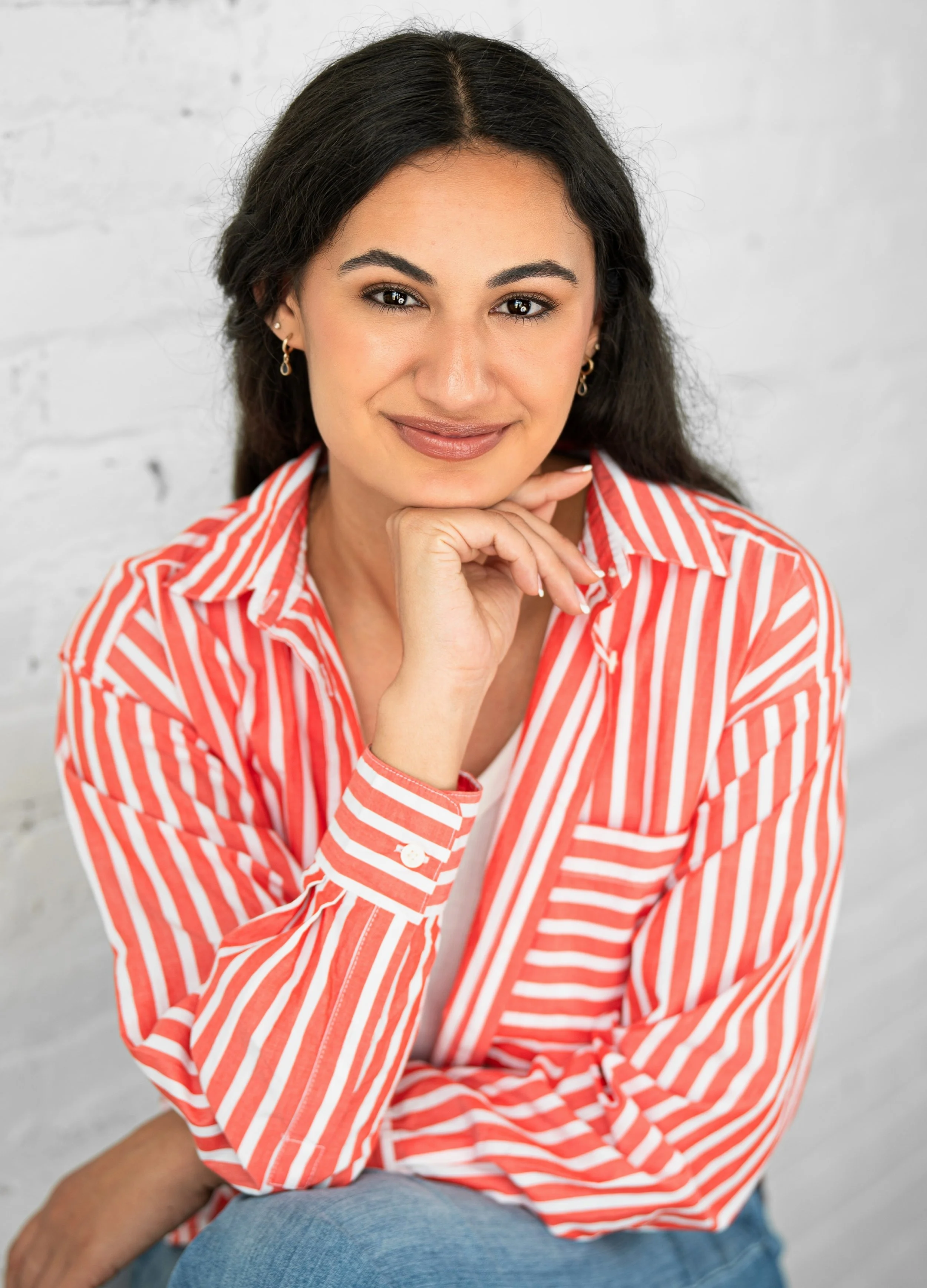 A woman with dark wavy hair, wearing a red and white striped shirt, sitting against a white brick wall, smiling and resting her chin on her hand.