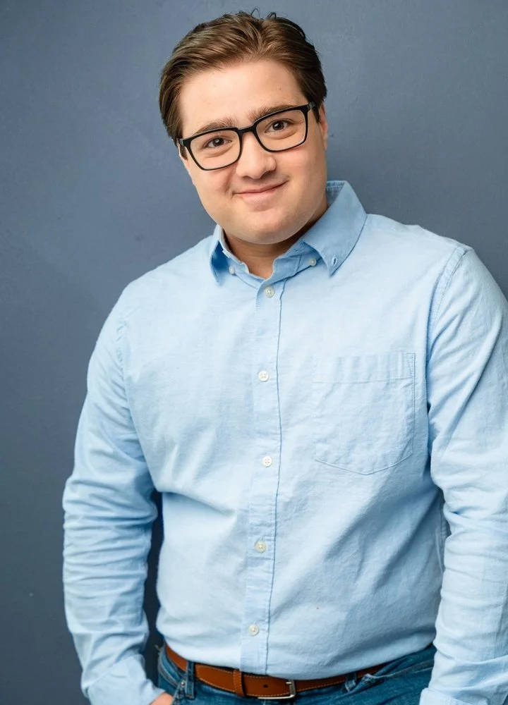 A young man with glasses, wearing a light blue button-up shirt, stands against a gray background, smiling at the camera.