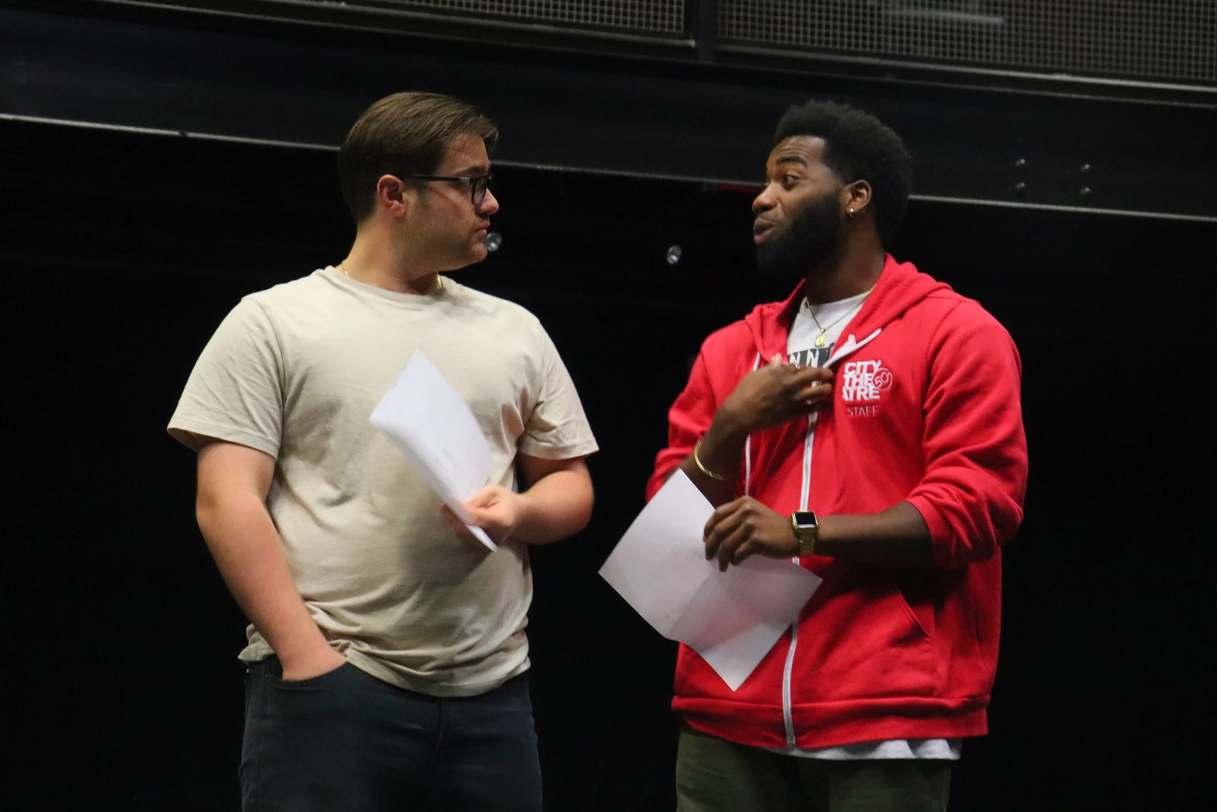 Two men engaged in conversation, holding papers, standing indoors against a dark background.