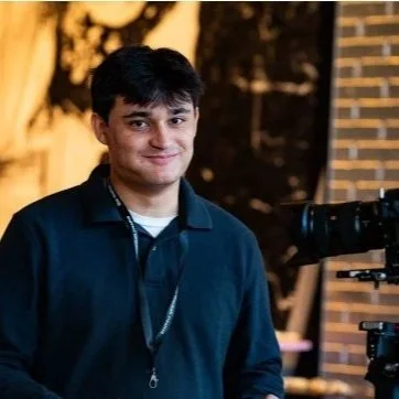 Young man with dark hair smiling, wearing a dark jacket, standing near a camera at an outdoor event during the evening.