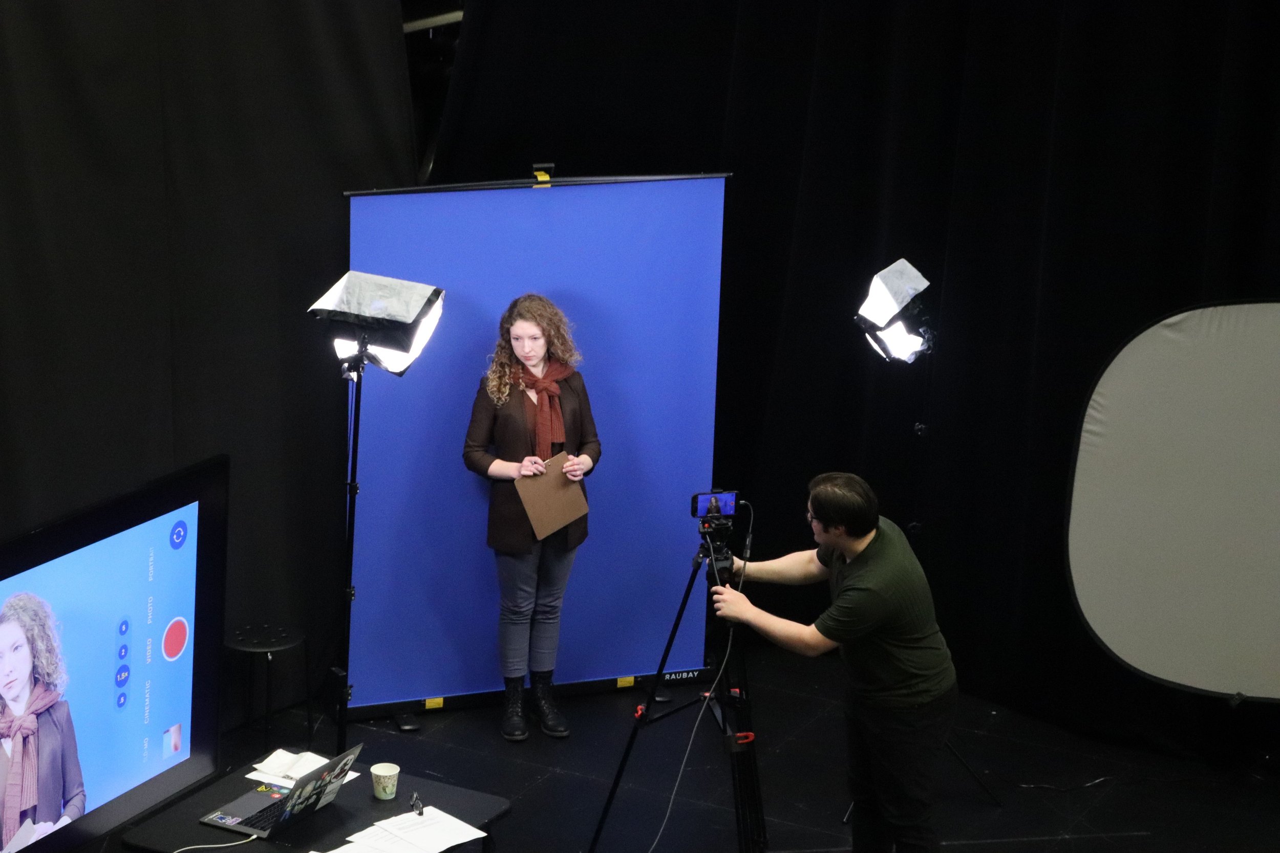 A woman standing in front of a blue screen during a recording or photo shoot with professional lighting, while a camera operator adjusts a camera on a tripod and a large display screen shows her image.