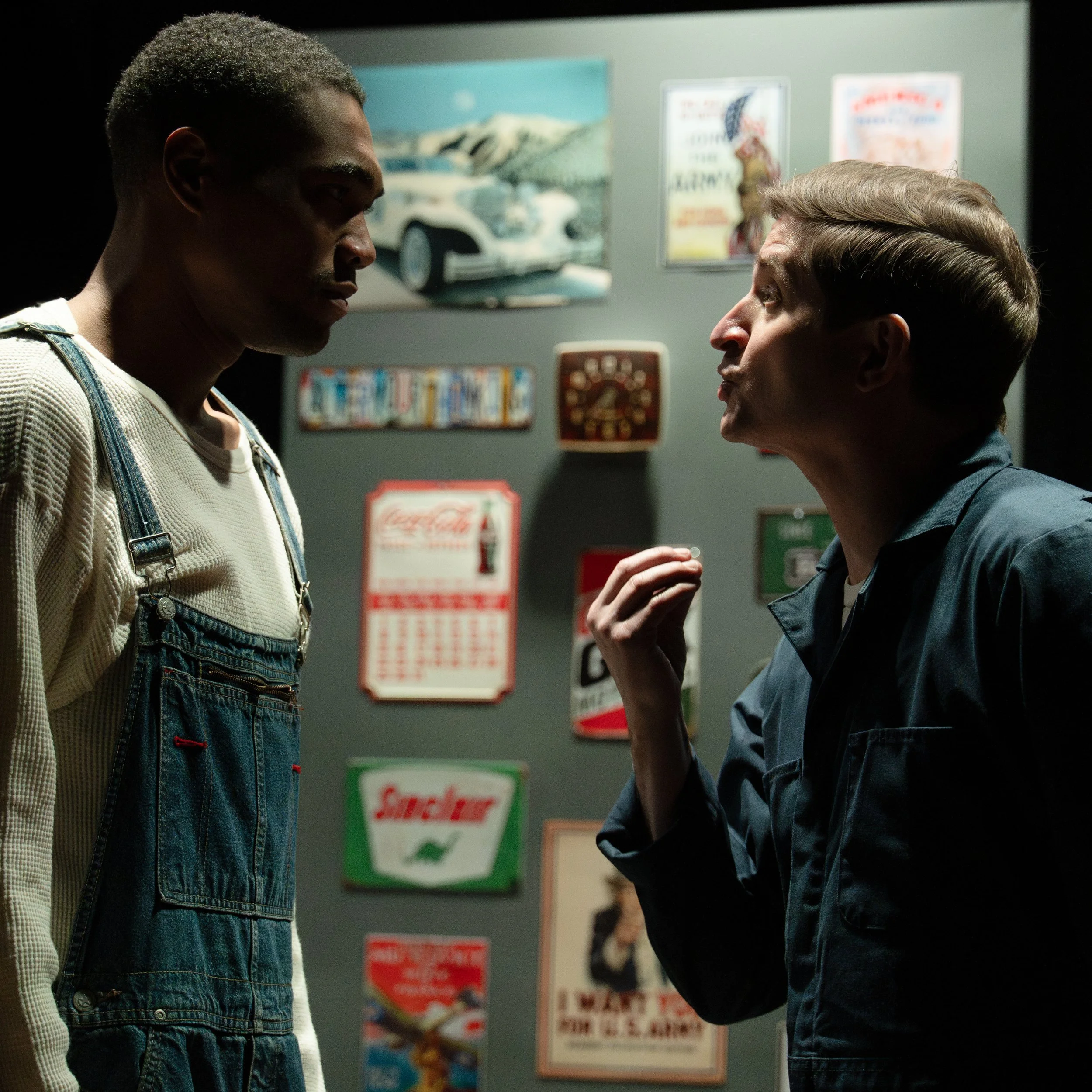Two men are facing each other closely in a dimly lit room, appearing to be in an intense conversation. The background shows vending machines and posters with bright colors.