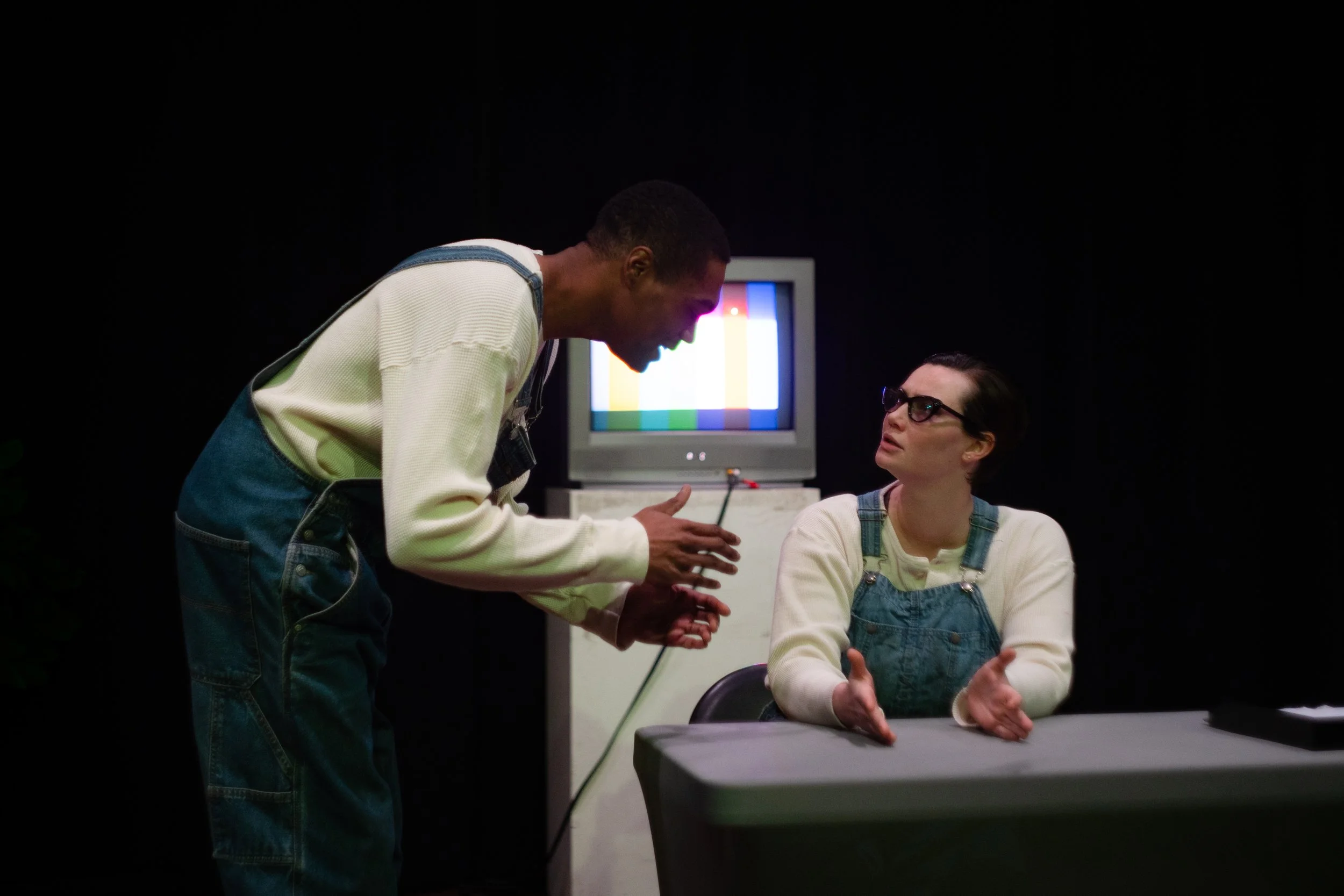Two people in a dark room with a television. The person on the left is standing and leaning toward the seated person, who is wearing glasses and overalls. The seated person looks up at the standing person with a serious expression.