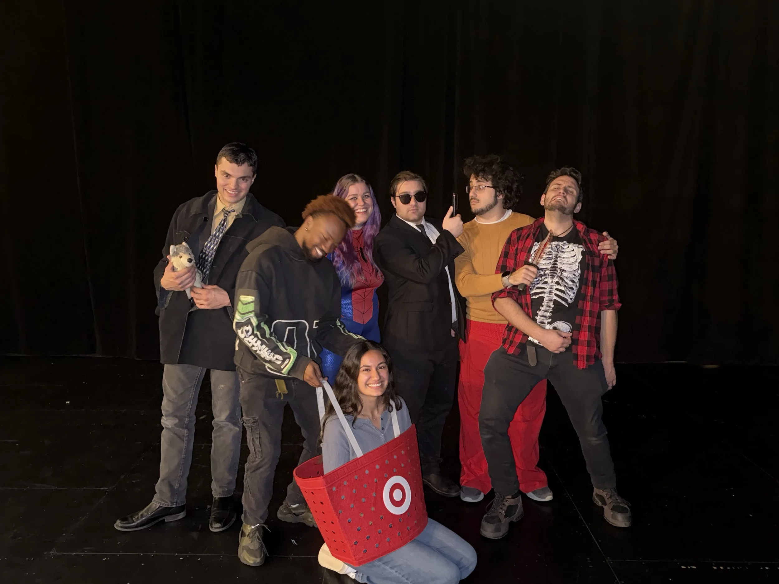 Group of people posing together, some smiling, one person holding a small stuffed animal, others with sunglasses and casual clothing, standing in front of a black curtain.