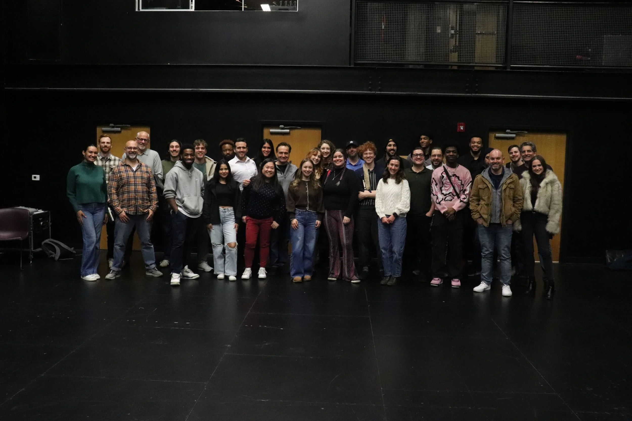 A large group of diverse people standing together in a dark indoor space, smiling at the camera.