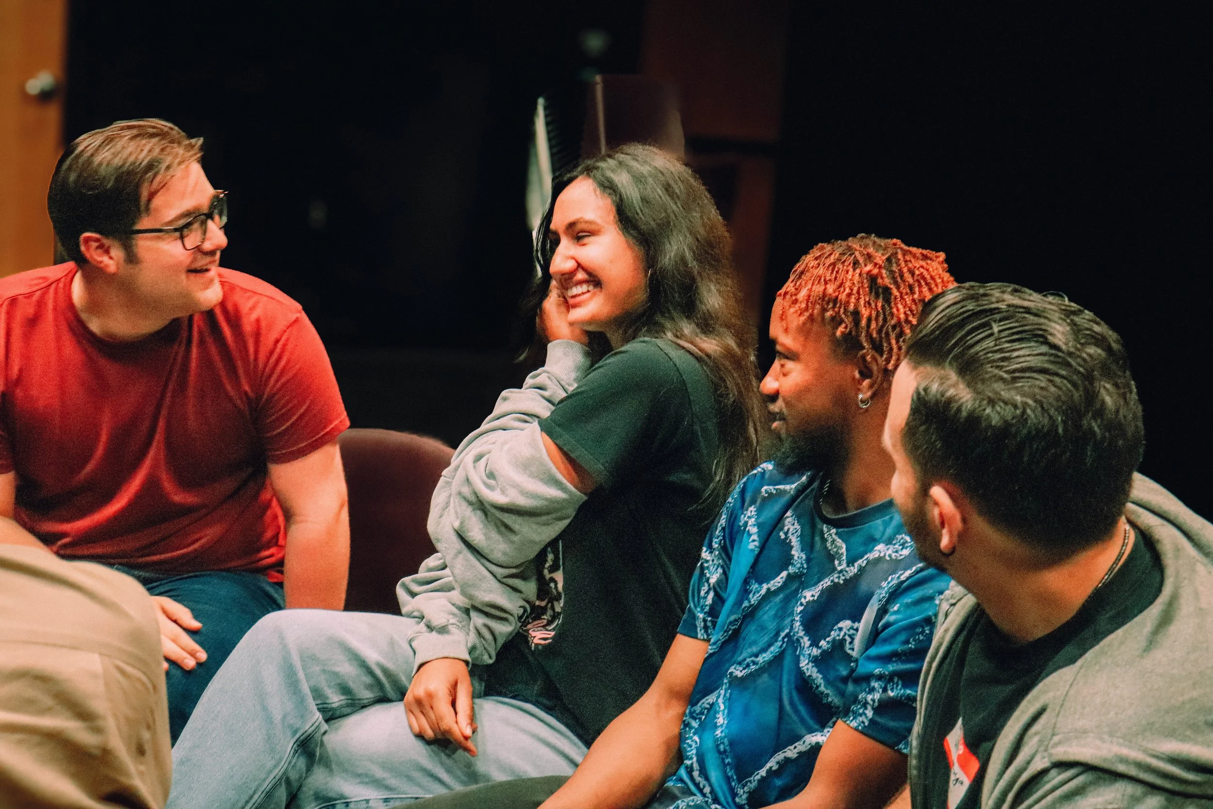 Group of diverse young adults sitting together, smiling and engaging in conversation in a social setting.