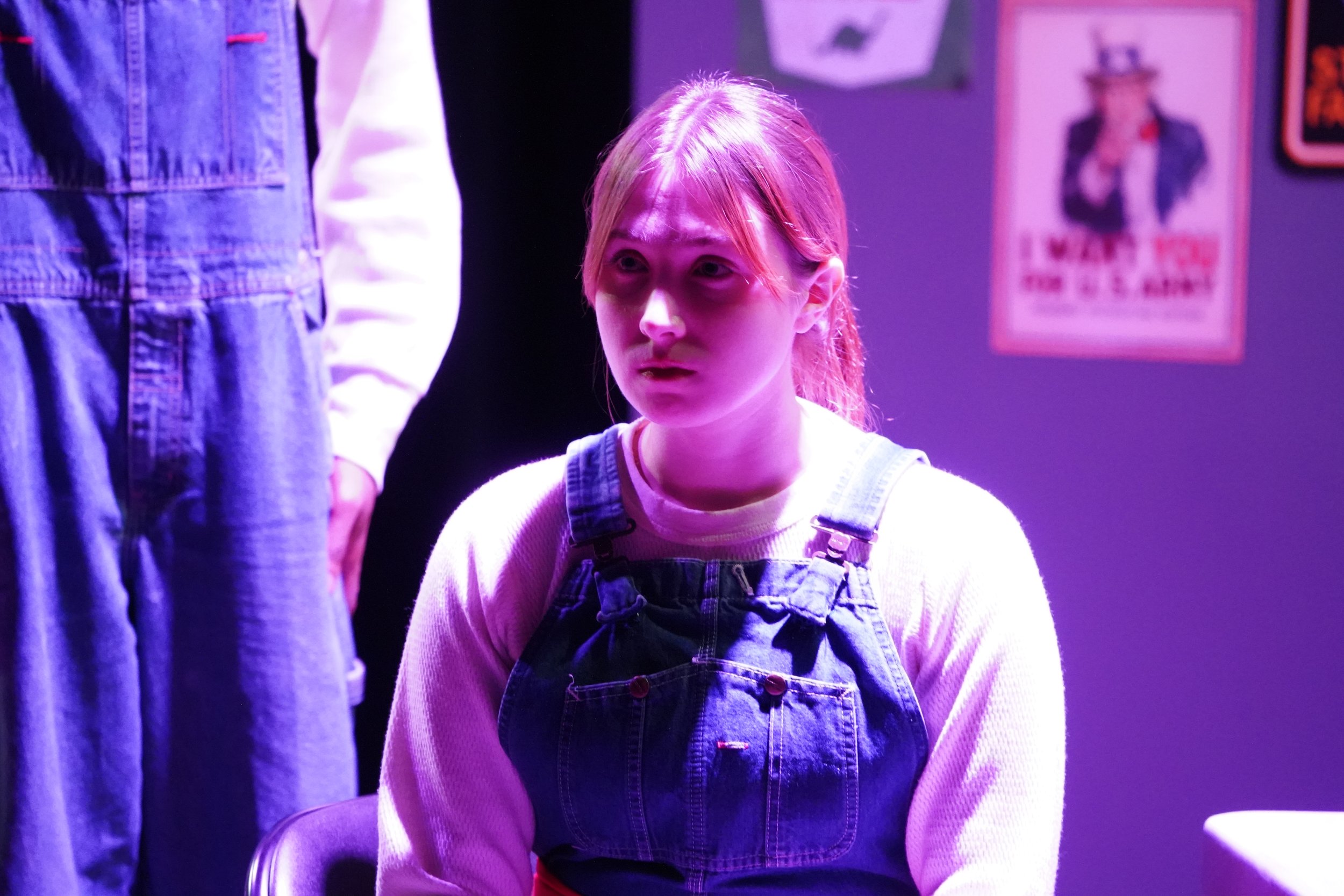 A young girl with pink hair and wearing overalls is sitting under purple lighting, looking at someone standing in front of her, whose torso and legs are partially visible.