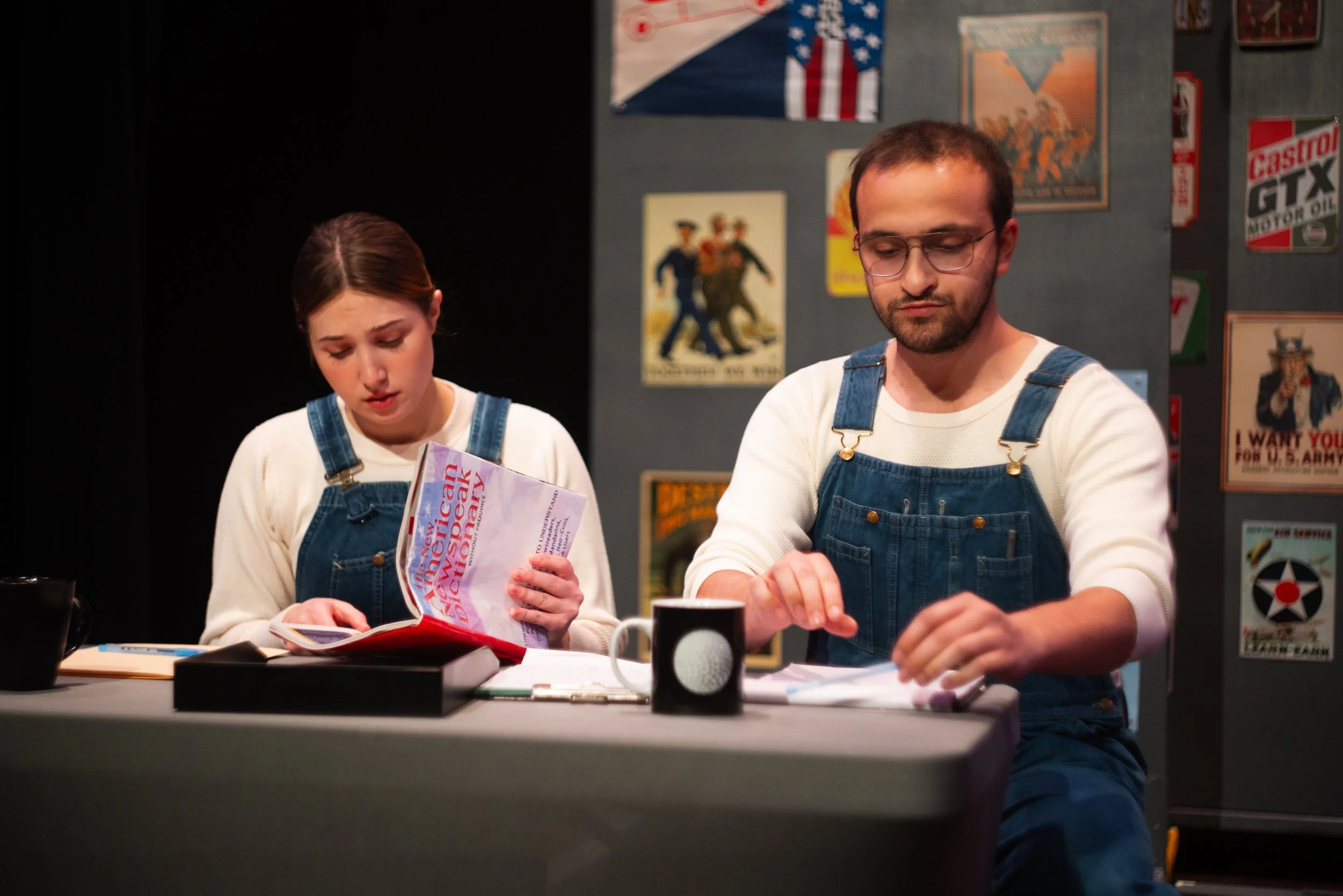 Two young adults, a woman with brown hair and a man with glasses, are sitting at a table wearing blue denim overalls and white shirts. They appear to be engaged in different activities; the woman is looking at a colorful magazine titled 'American History' while the man is focused on something in front of him. The backdrop has vintage posters and flags, including an American flag and related patriotic posters.