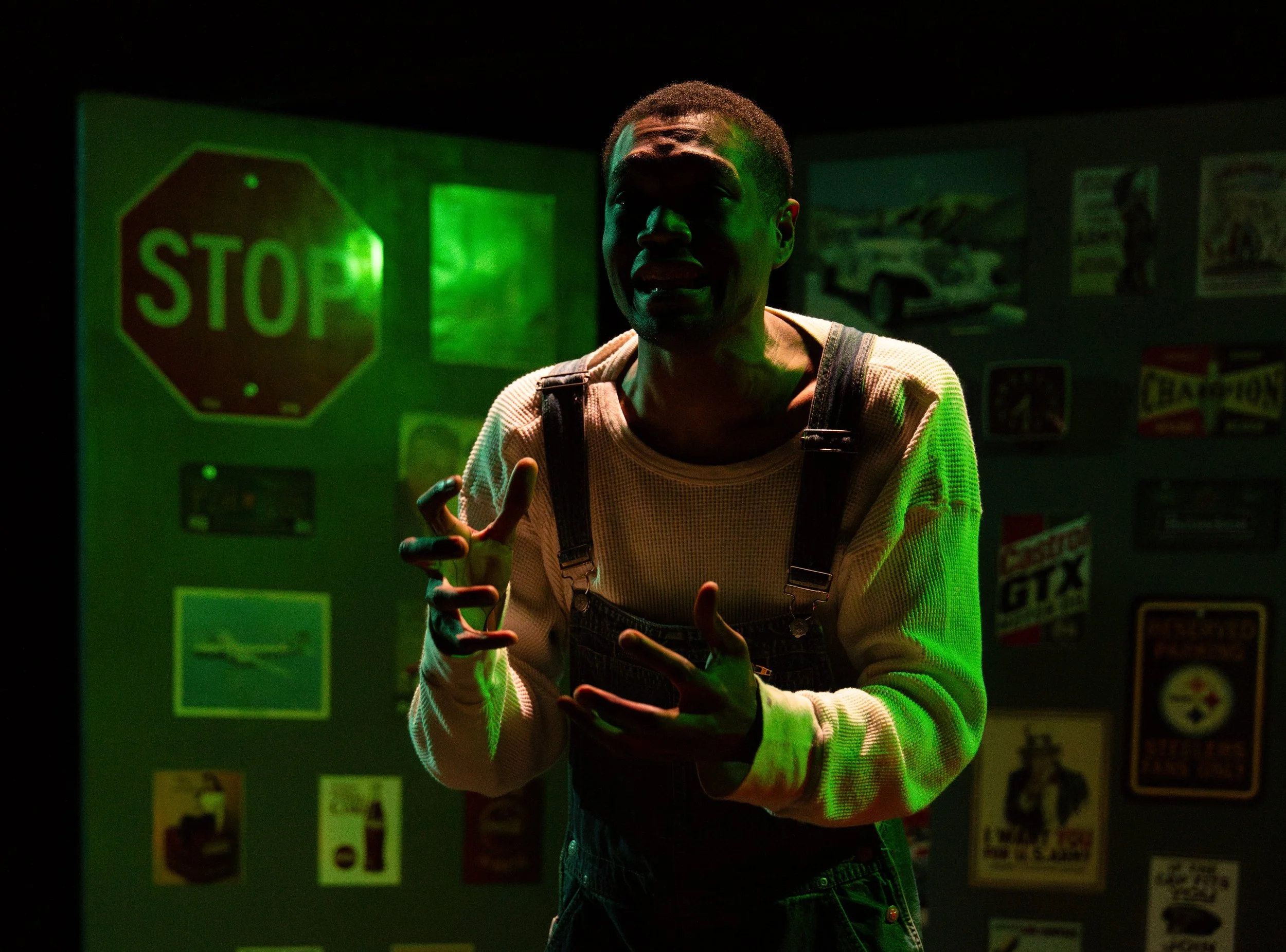 A young man with dark skin and short hair standing in a dimly lit room with green light, making a peace sign with both hands, and smiling with his tongue slightly out. There are signs on the wall behind him, including a large green stop sign.