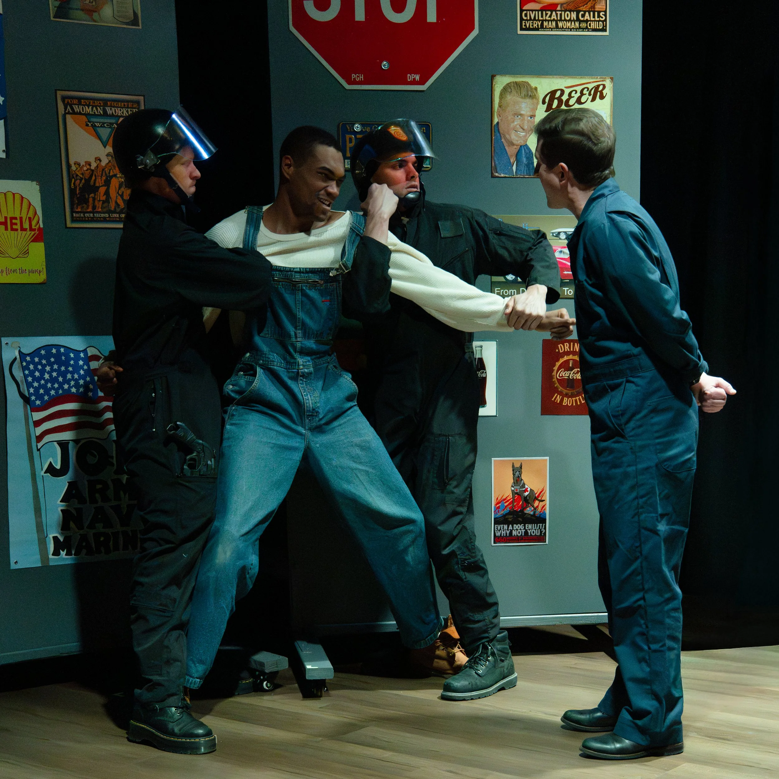 Several men are in a room with vintage posters on the wall. Two men are wearing helmets and holding hands, shaking hands with a third man. They appear to be engaging in a friendly handshake or greeting.