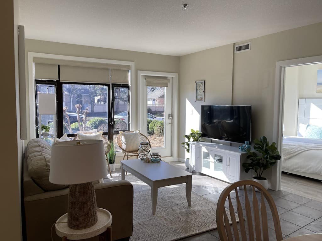 Living room with large windows, beige sofa, white coffee table, and flat-screen TV on a white stand, with a bedroom visible through a doorway.