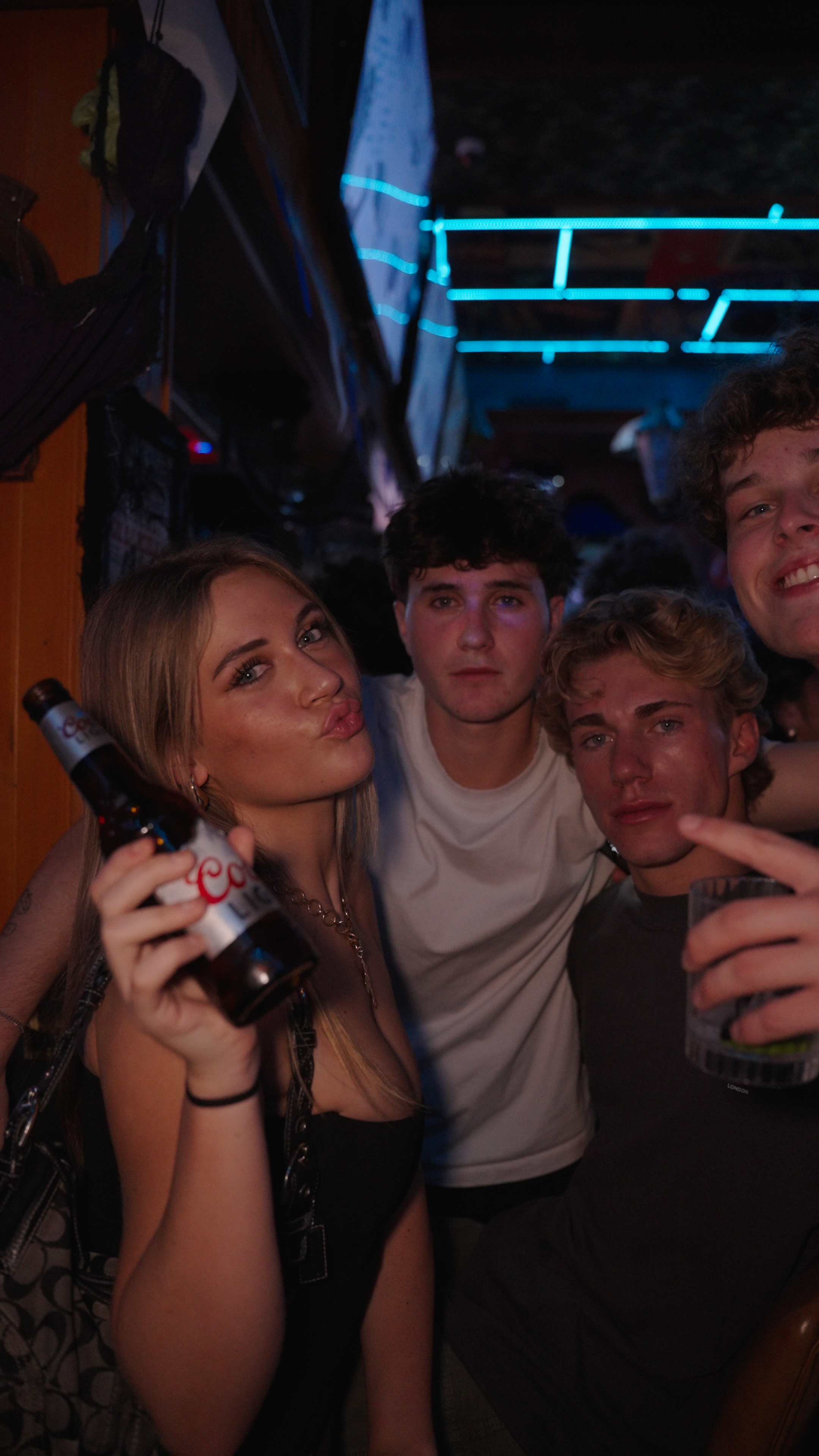 Group of young adults at a party, posing for a photo with drinks in hand, in a dimly lit environment with neon blue lights in the background.