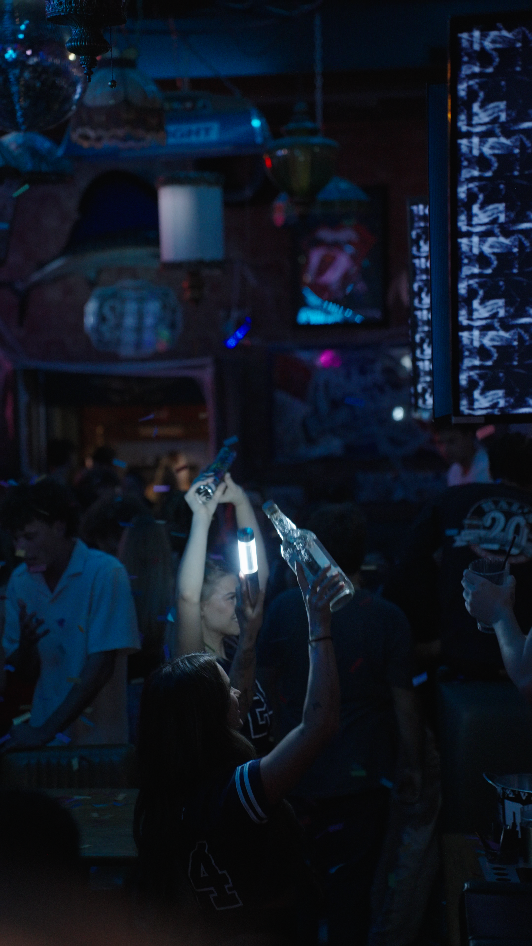 People dancing and holding bottles in a nightclub with dark lighting, a large screen, and colorful lights.