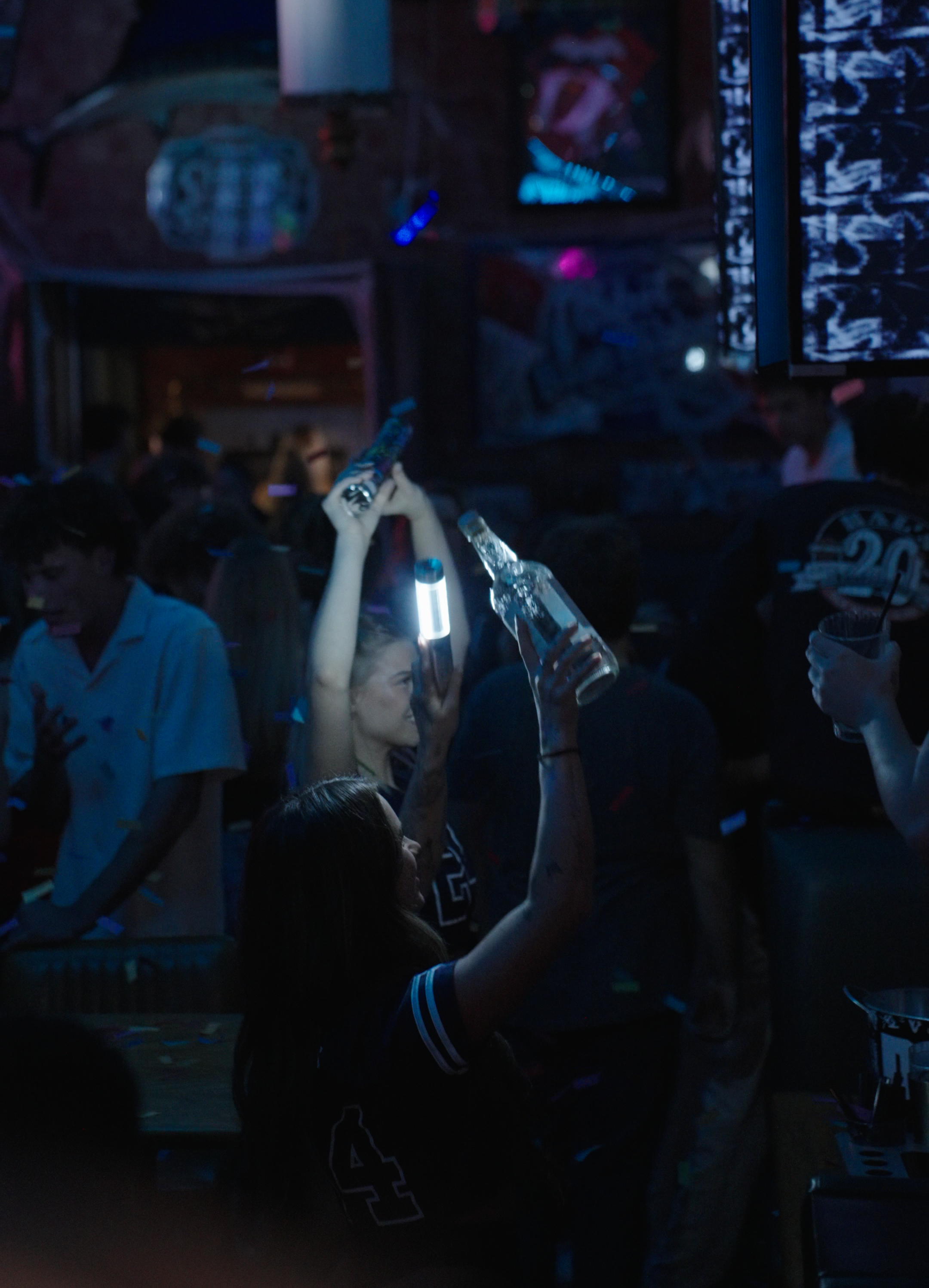 People in a nightclub holding drinks, surrounded by blue and pink lighting, with a digital screen and confetti in the air.