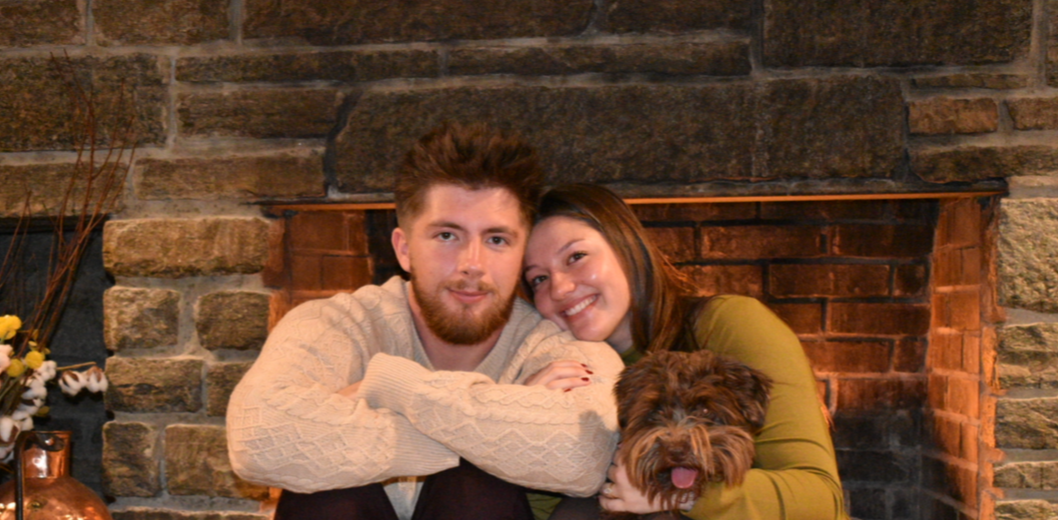 A couple sitting in front of a brick fireplace with their dog, smiling.