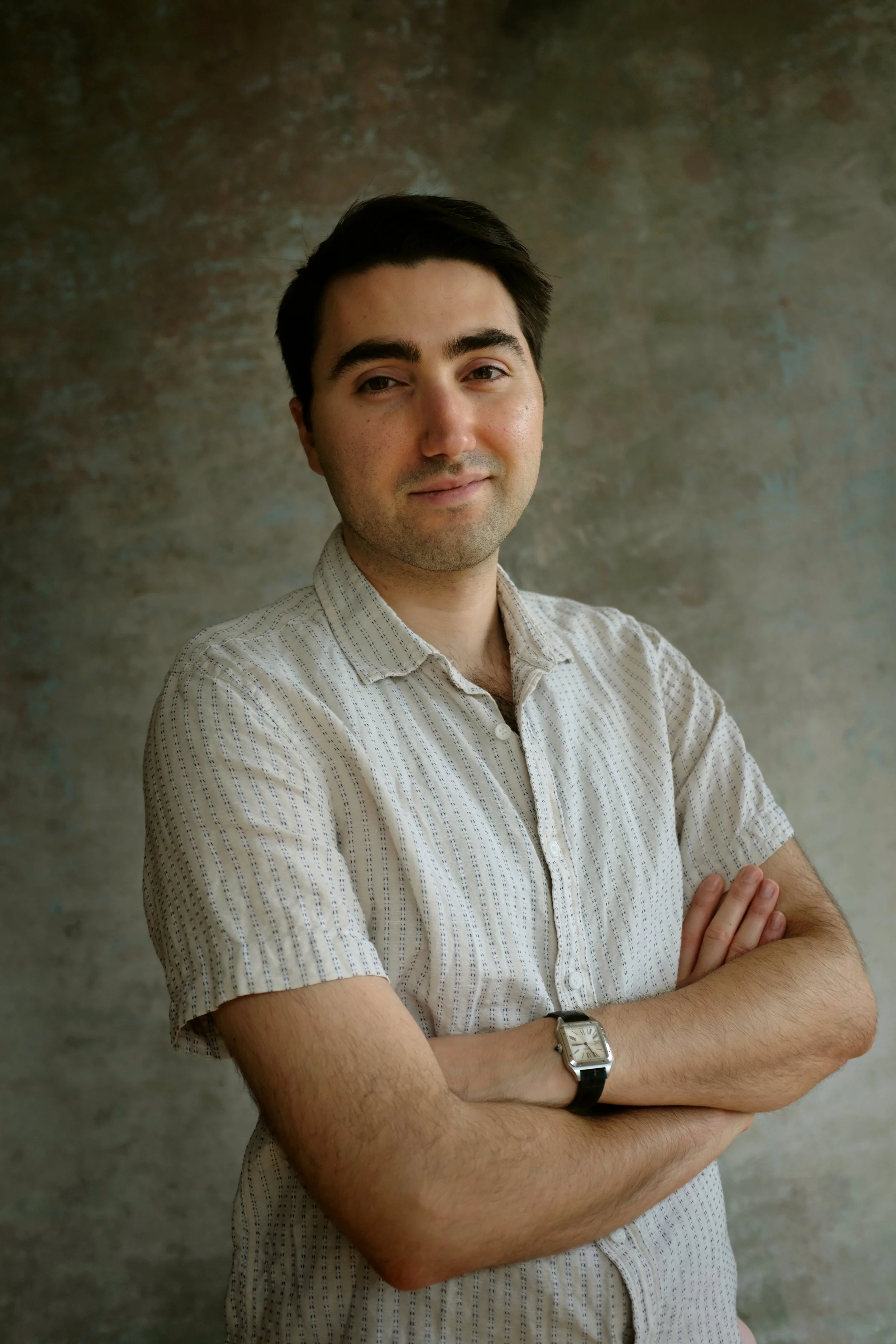 A young man with dark hair, wearing a short-sleeved, light-colored, striped button-up shirt, crosses his arms, and wears a watch on his left wrist, standing in front of a textured, multicolored background.