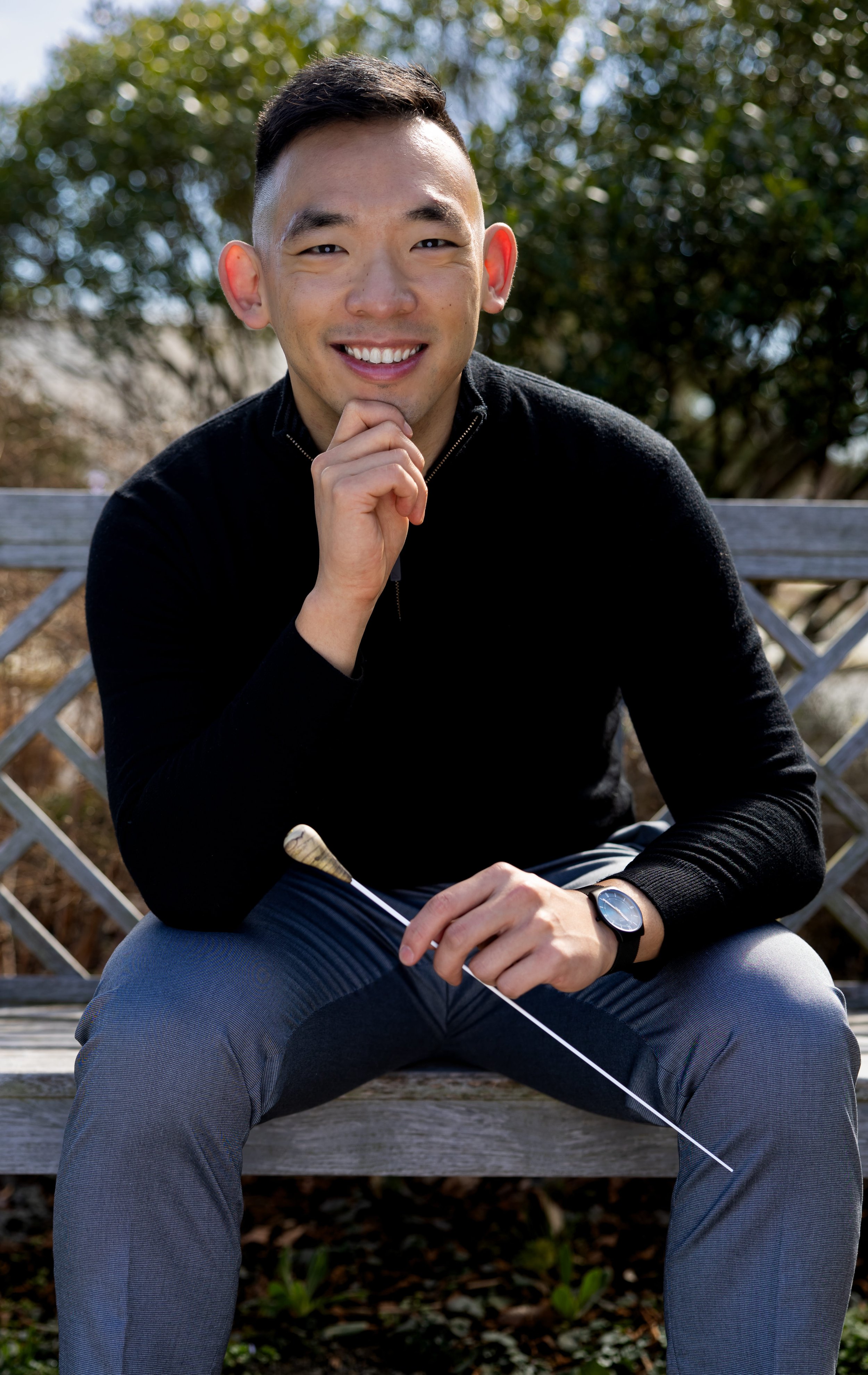 A smiling young Asian man sitting on a wooden bench outdoors, holding a golf club, wearing a black long-sleeve shirt, gray pants, and a black watch.