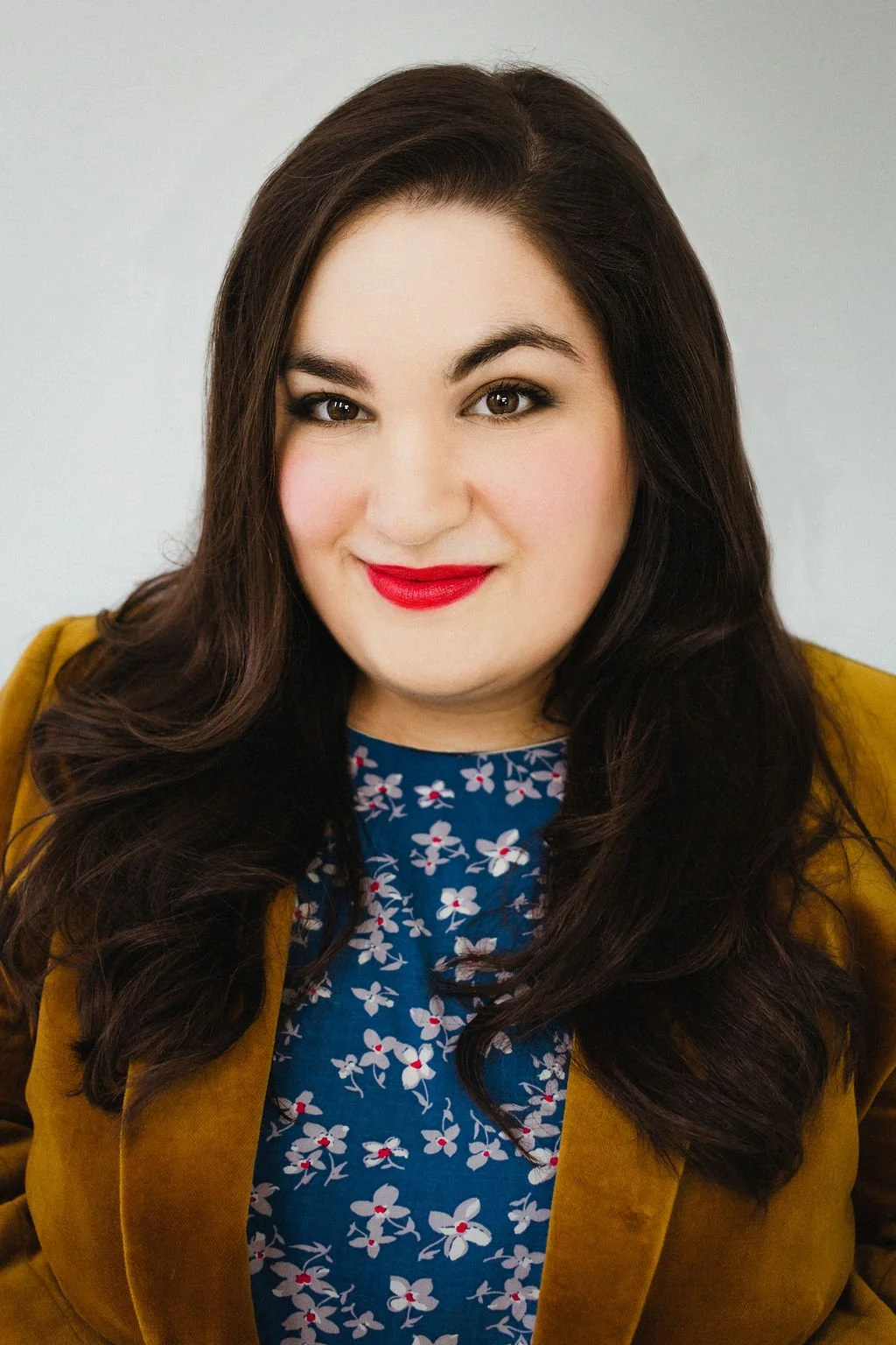 A woman with long, dark brown hair and red lipstick, wearing a blue floral top and a brown jacket, smiling at the camera.