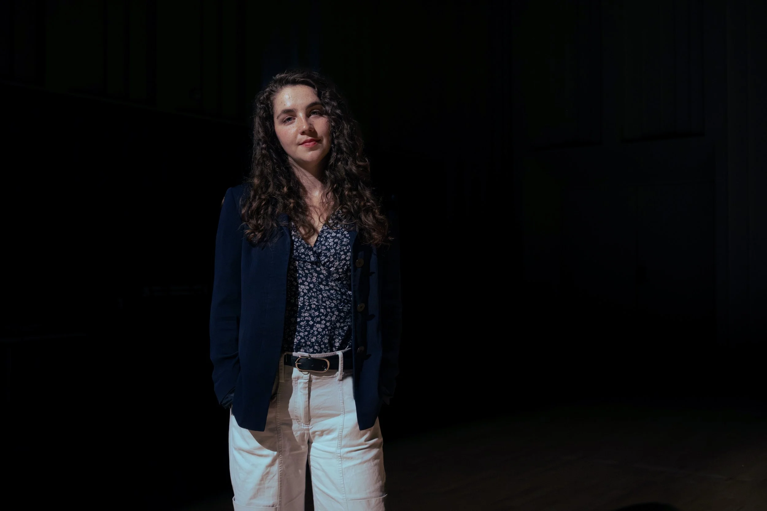 A young woman with long curly brown hair and light skin stands in a dimly lit room with dark walls, wearing a navy blazer, a floral blouse, and beige pants.