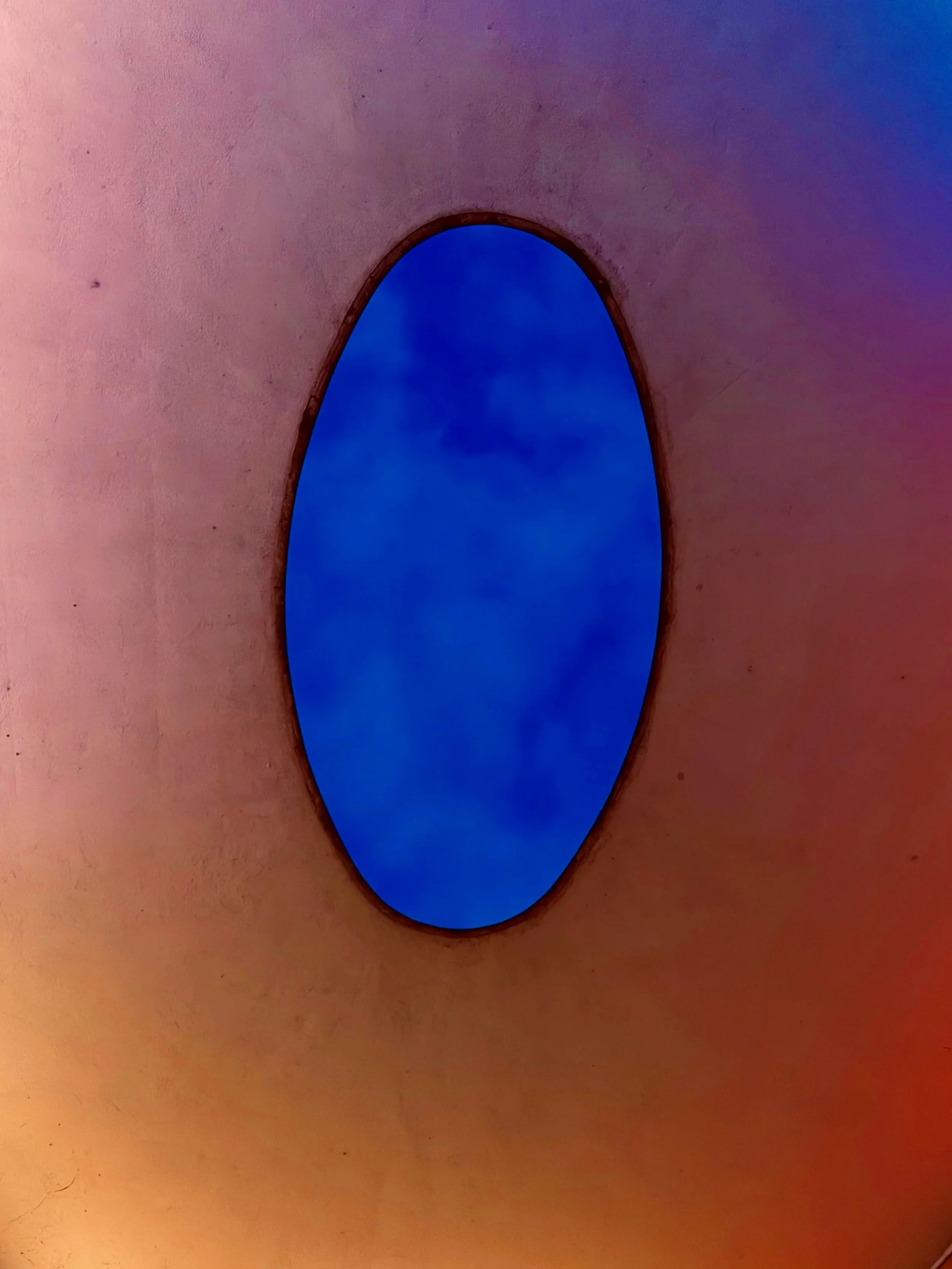 Close-up view of an oval window set into a beige-colored wall with a blue sky and clouds visible through the window.