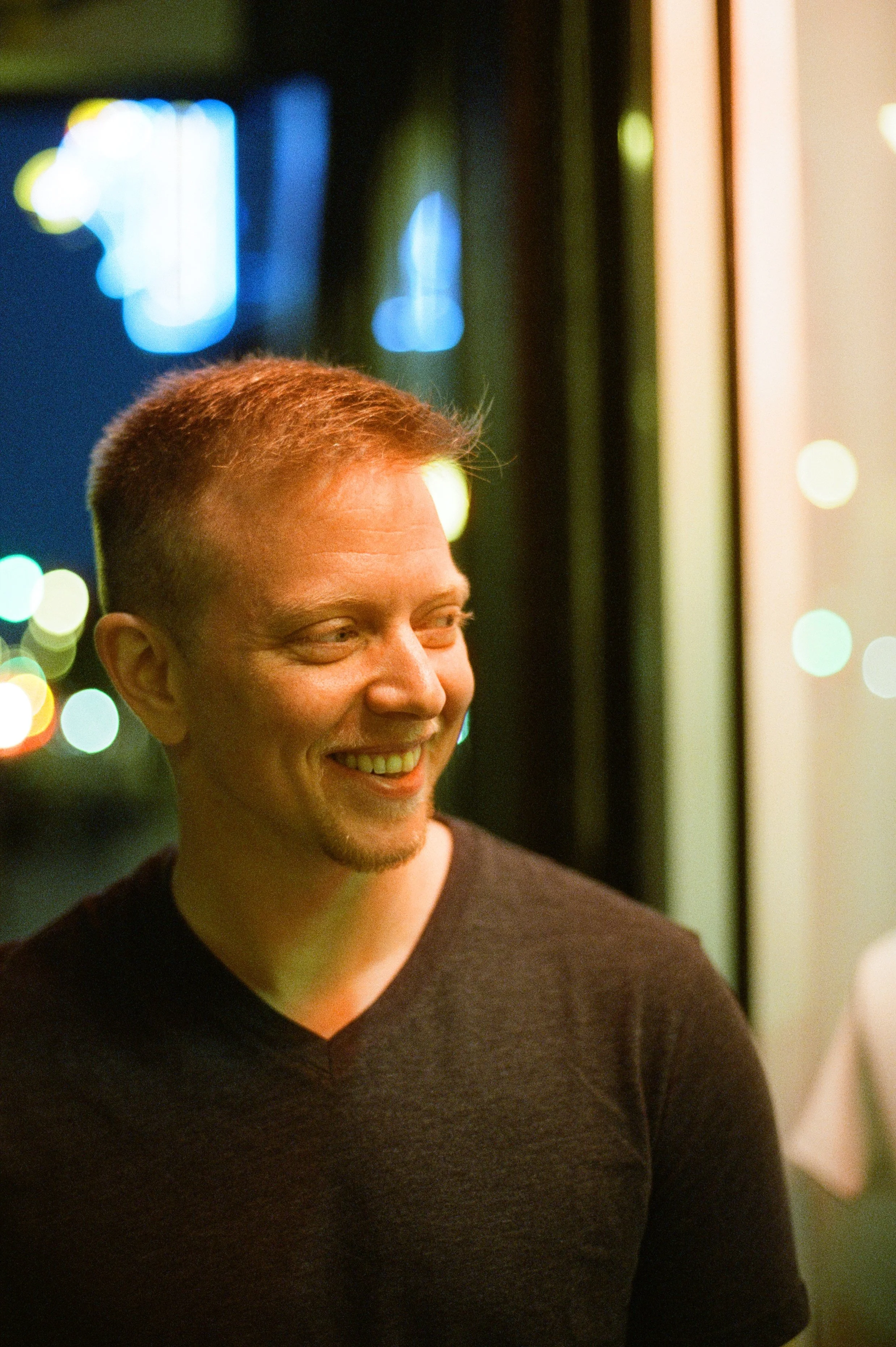 A smiling young man with short red hair, wearing a black t-shirt, standing indoors at night with blurred colorful city lights in the background.