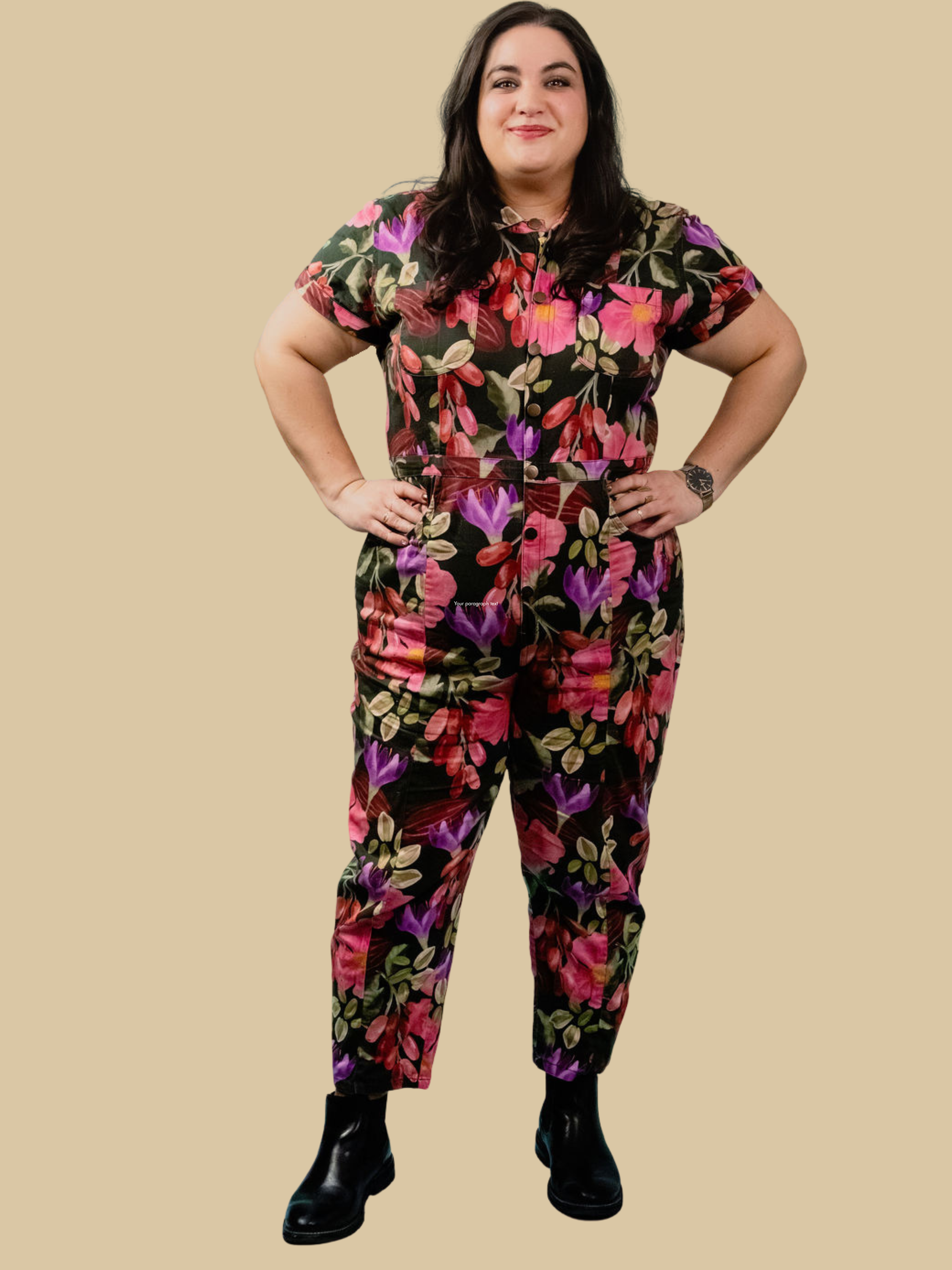 Woman with long dark hair wearing a floral jumpsuit with pink and purple flowers, and red fruit, standing with hands on hips against a beige background.