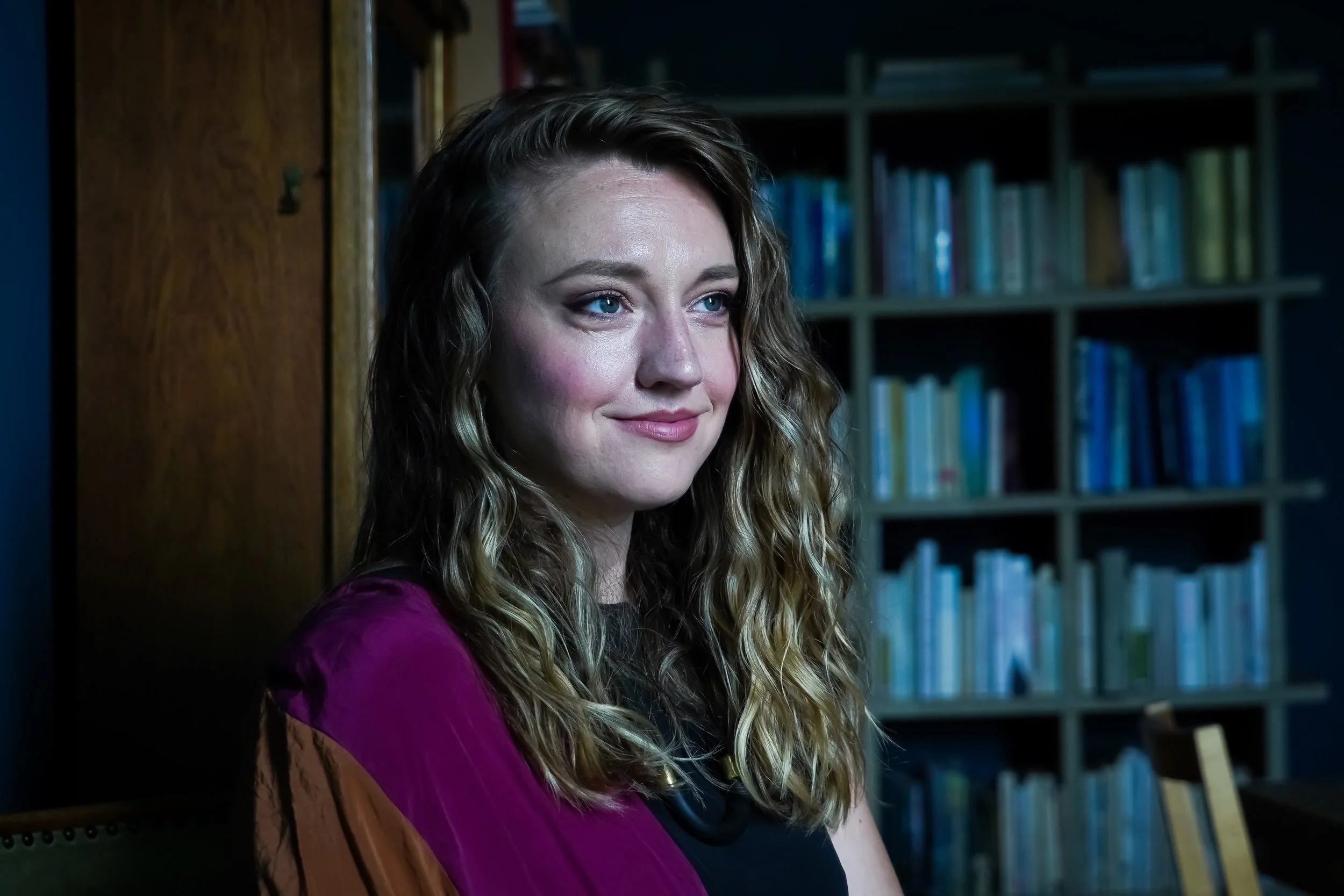 A young woman with long, wavy, light brown hair and blue eyes smiling softly, sitting indoors near a wooden bookshelf filled with colorful books.