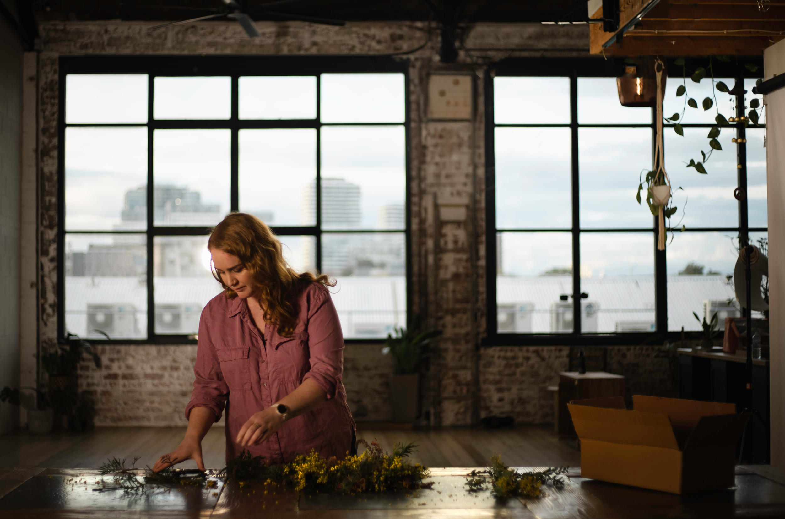 A woman in a pink shirt arranging flowers or plants on a wooden table in a spacious room with large windows showing city buildings outside.