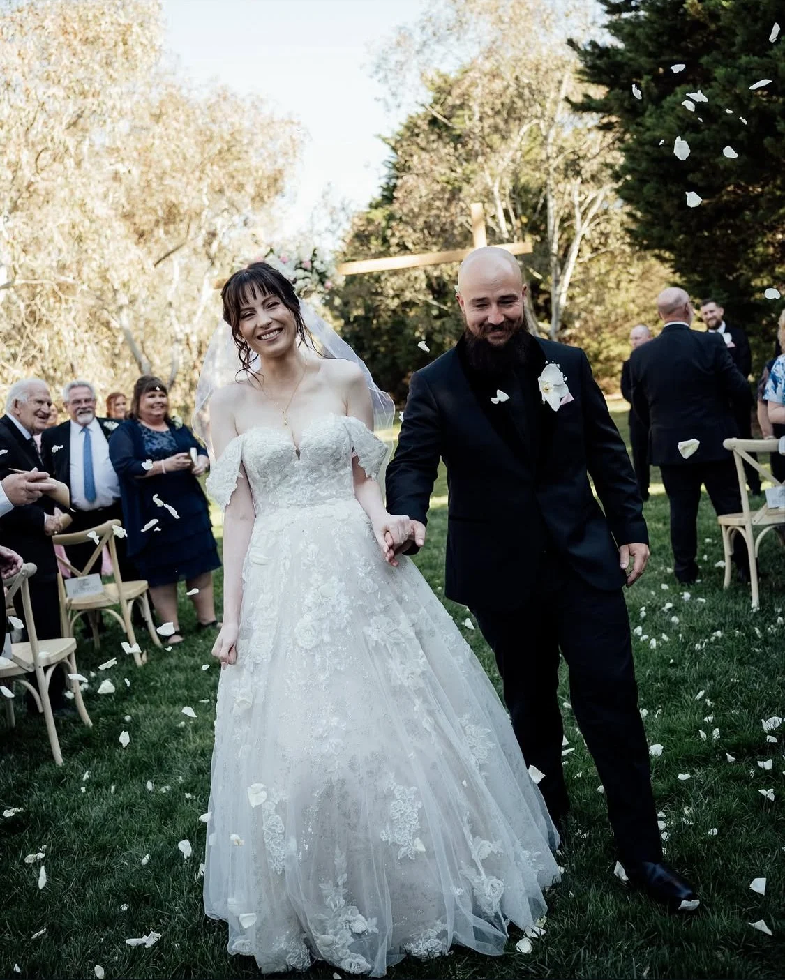 A bride and groom walking outdoors after their wedding ceremony, surrounded by smiling guests, with a cross in the background and flower petals falling.