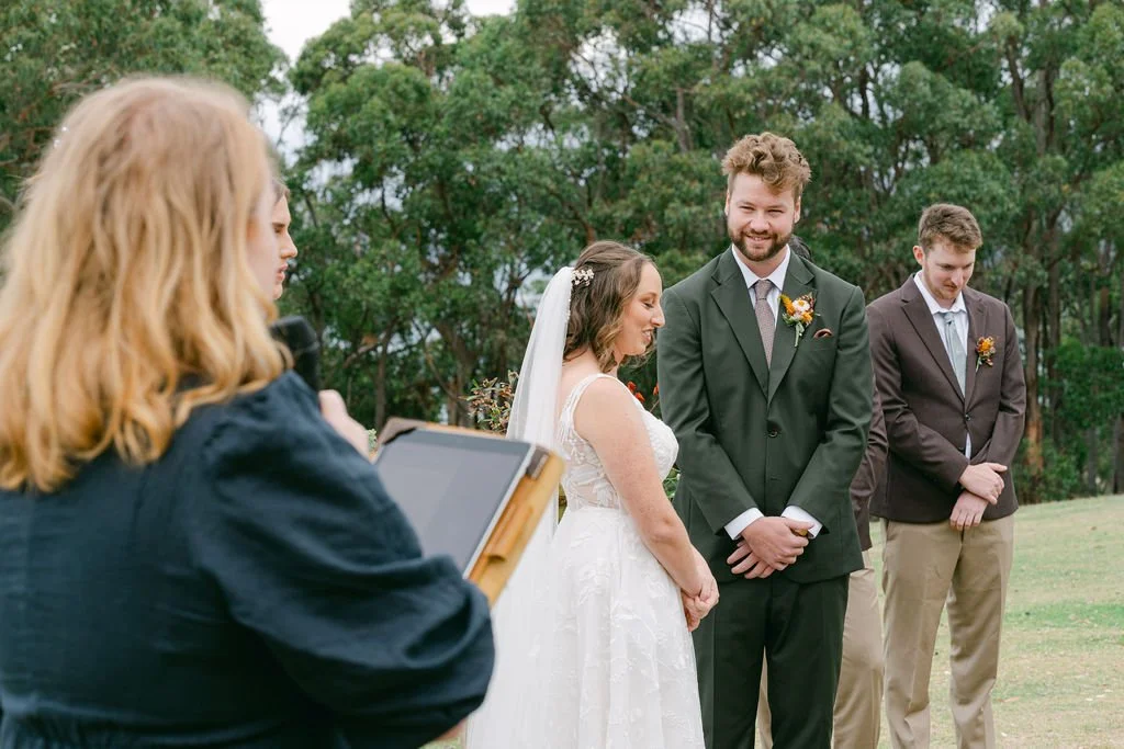 A bride and groom exchanging vows during an outdoor wedding ceremony in a green park with bridesmaids and groomsmen, with a female officiant holding a tablet or book.