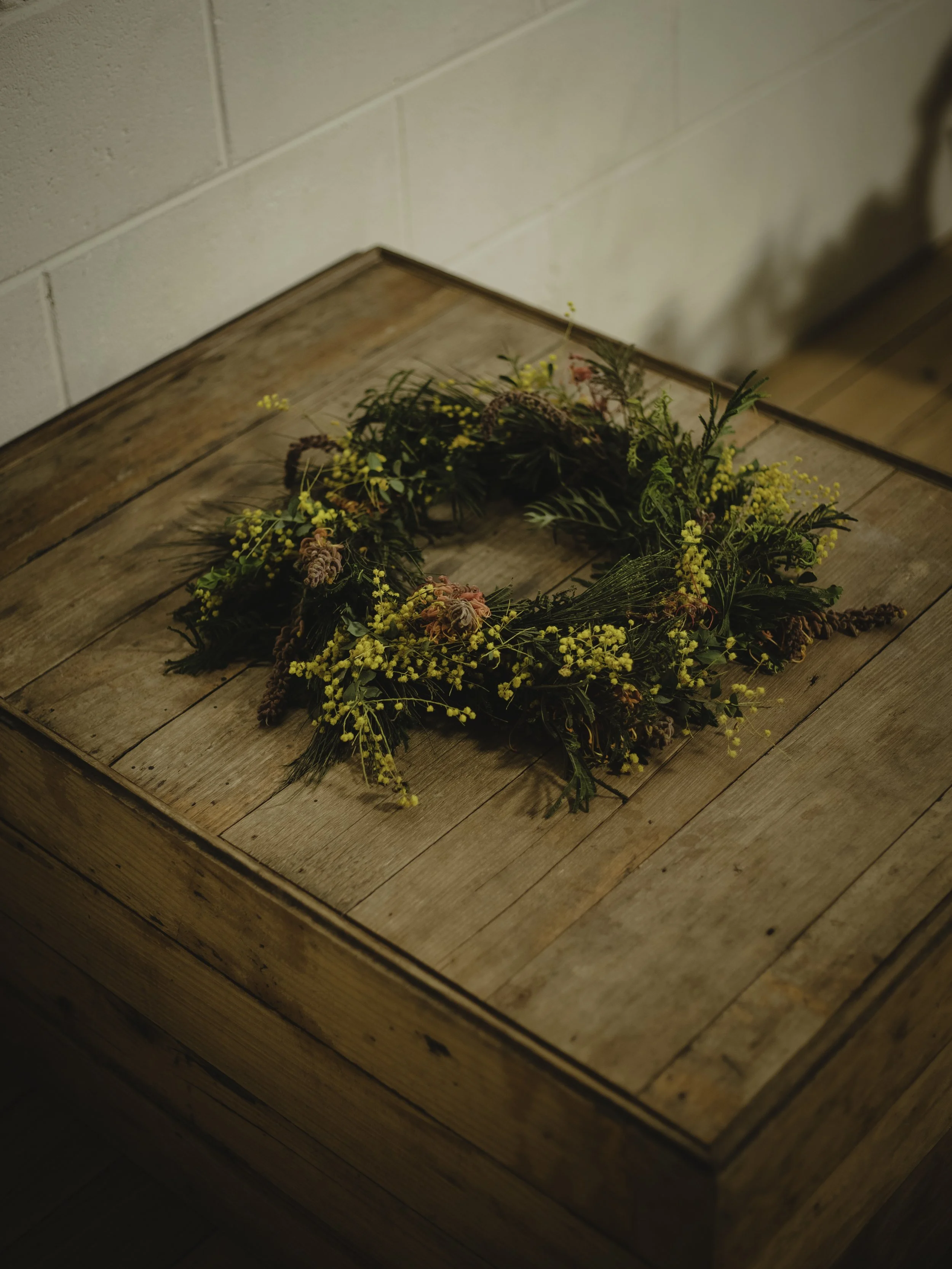 A decorative wreath made of yellow flowers, green leaves, and some pink accents on a wooden table.