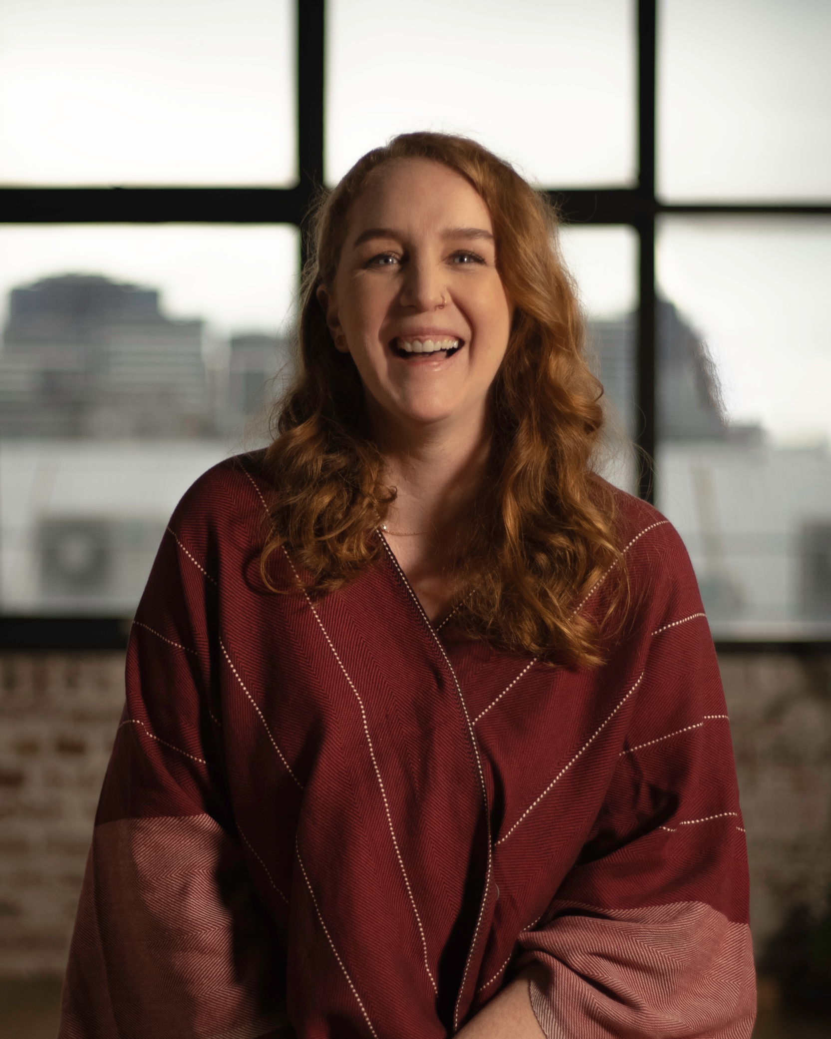 A young woman with curly red hair smiling inside a building with large window panes, cityscape in the background.
