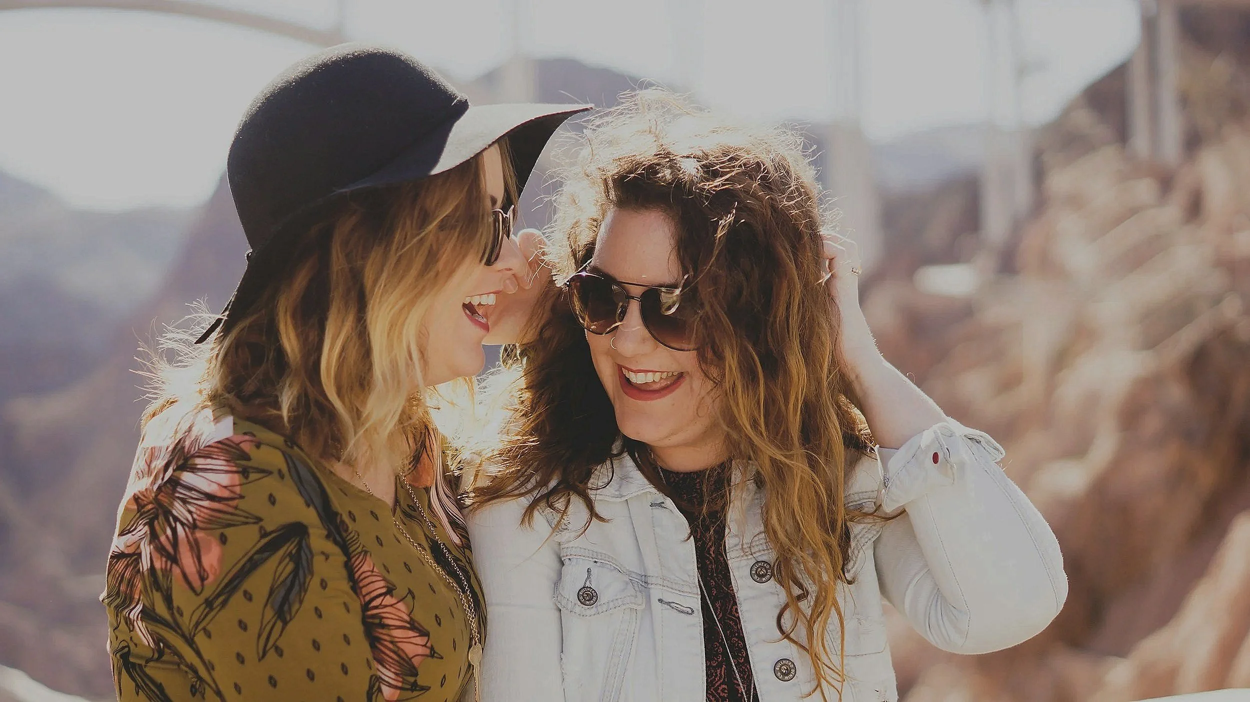 Two women smiling and talking, one wearing a black hat and sunglasses, the other wearing a white jacket and sunglasses, outdoors with a mountainous background.