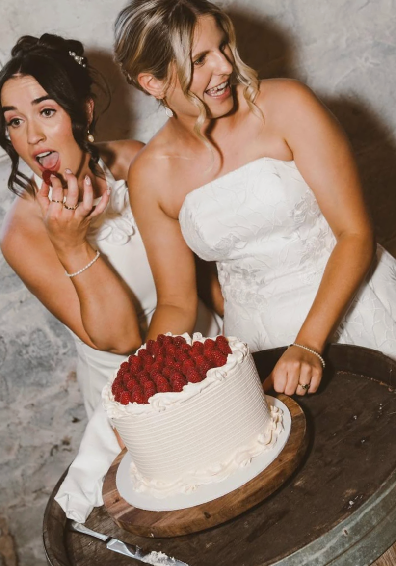 Two women in wedding dresses celebrating a wedding, with one holding a cake topped with strawberries.