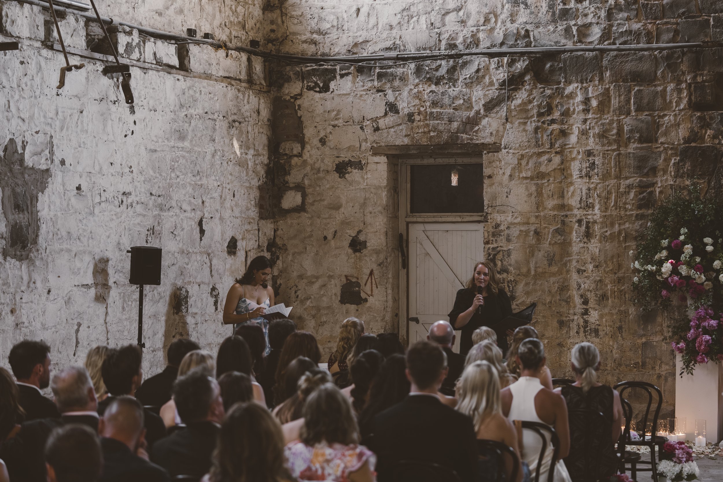A woman stands speaking into a microphone at a wedding or event in a rustic stone-walled venue, with a large bouquet of pink and white flowers on a pedestal to her left, and an audience seated listening.