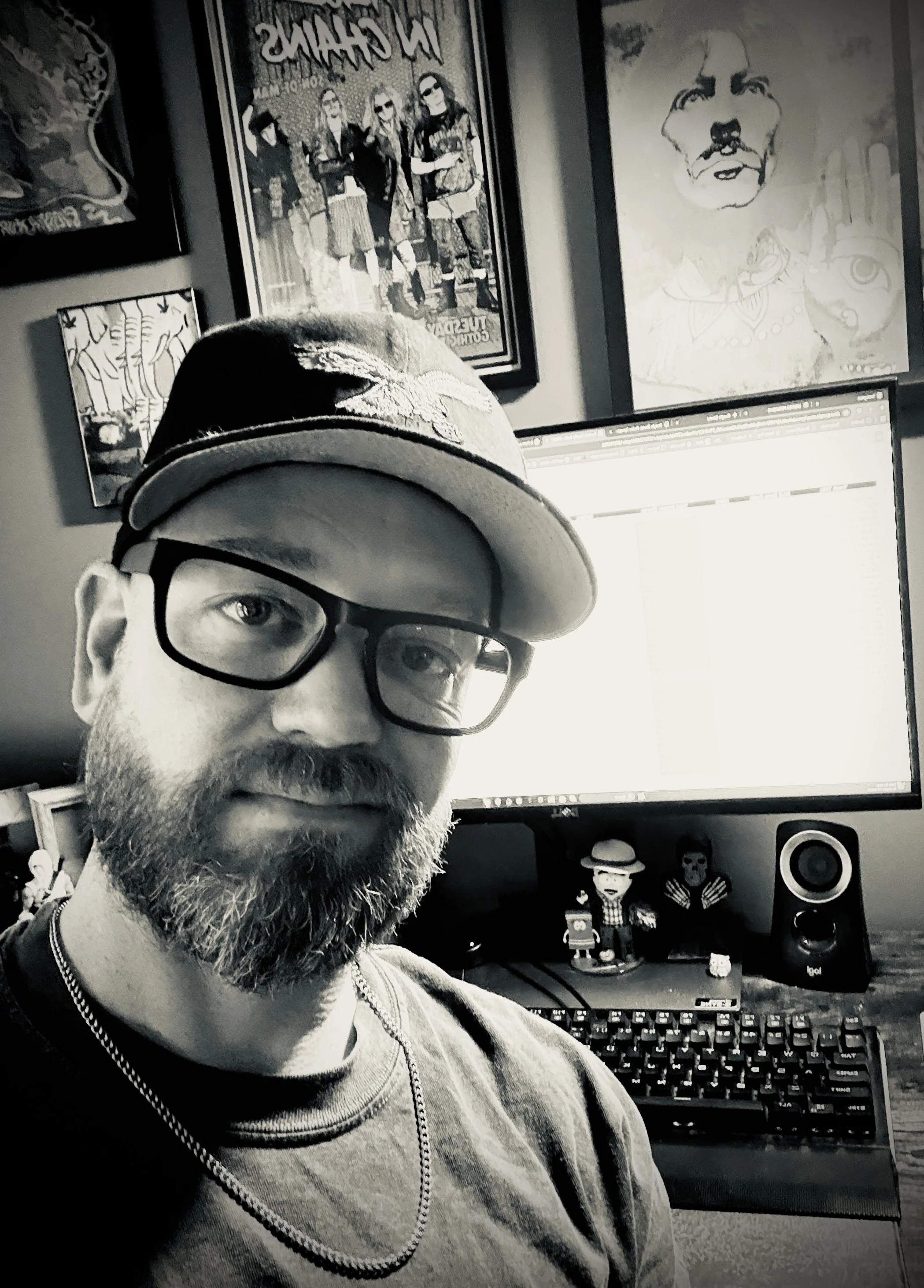 A man with glasses and a beard wearing a baseball cap and a necklace, sitting at a desk with a computer monitor, keyboard, and speakers. The wall behind him displays framed posters and artwork.