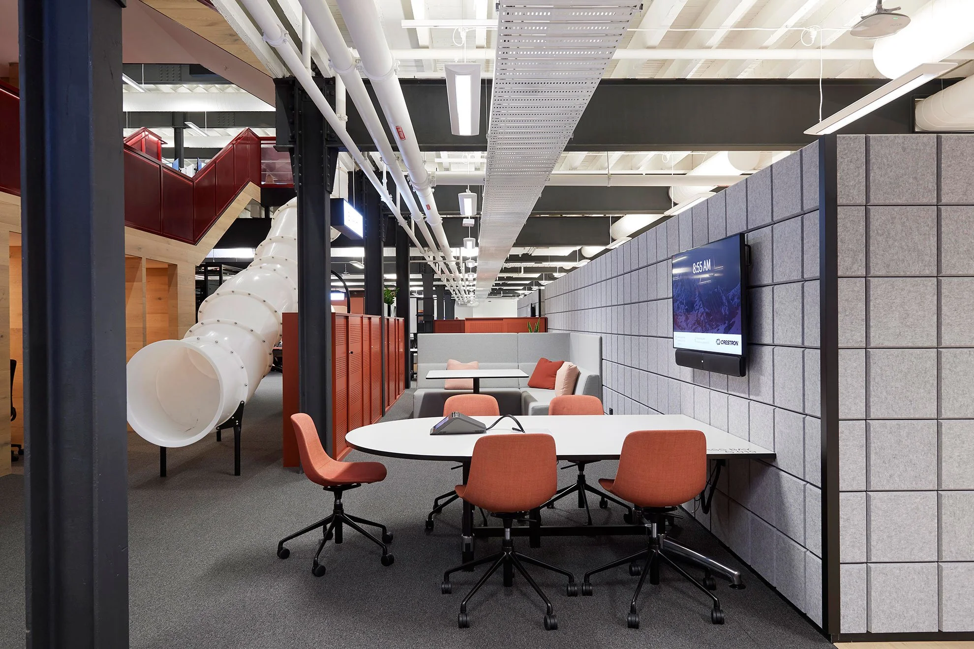 Modern office space with tables and orange chairs, gray cushioned seating area, mounted digital clock on wall, and a large white slide leading to upper level.