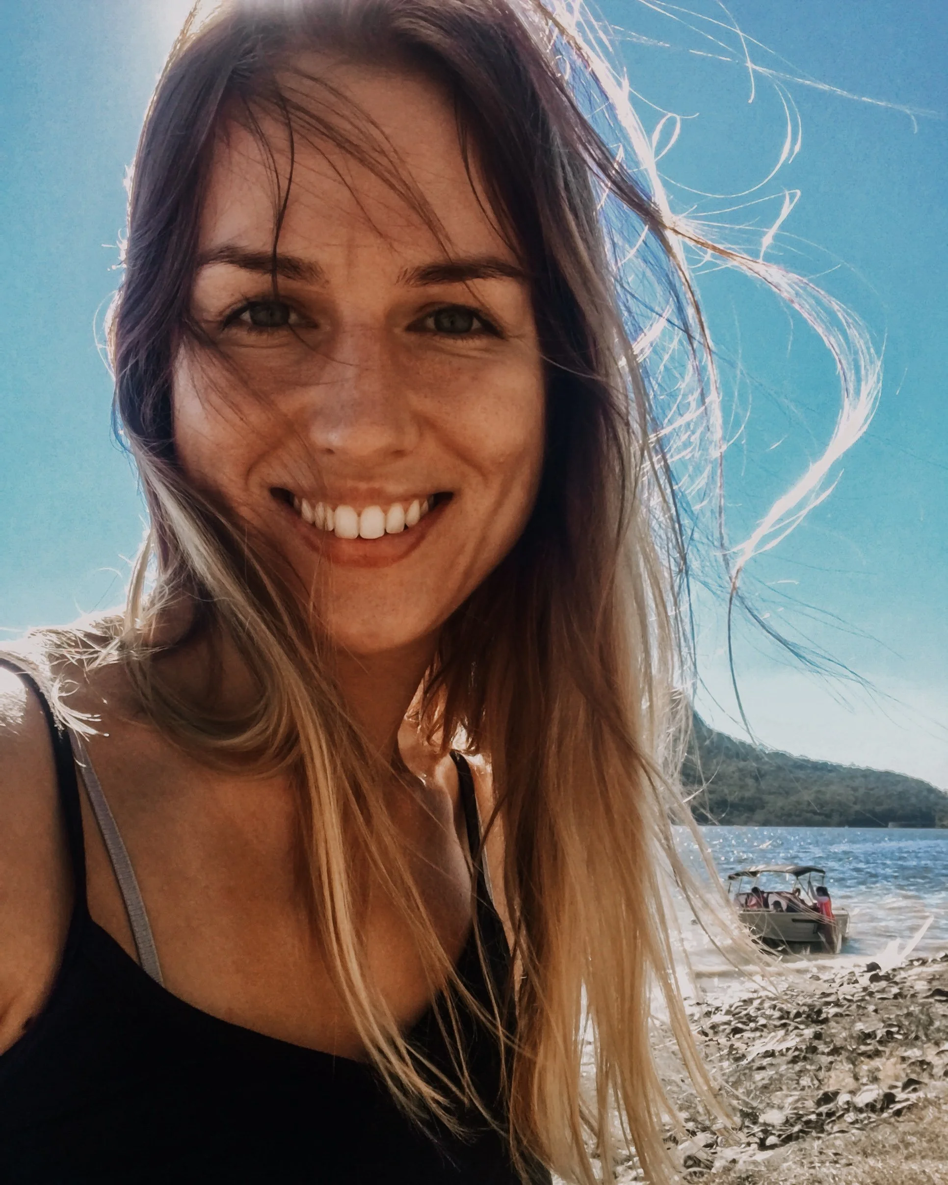 A smiling woman with long hair at the beach, with a boat and water in the background.