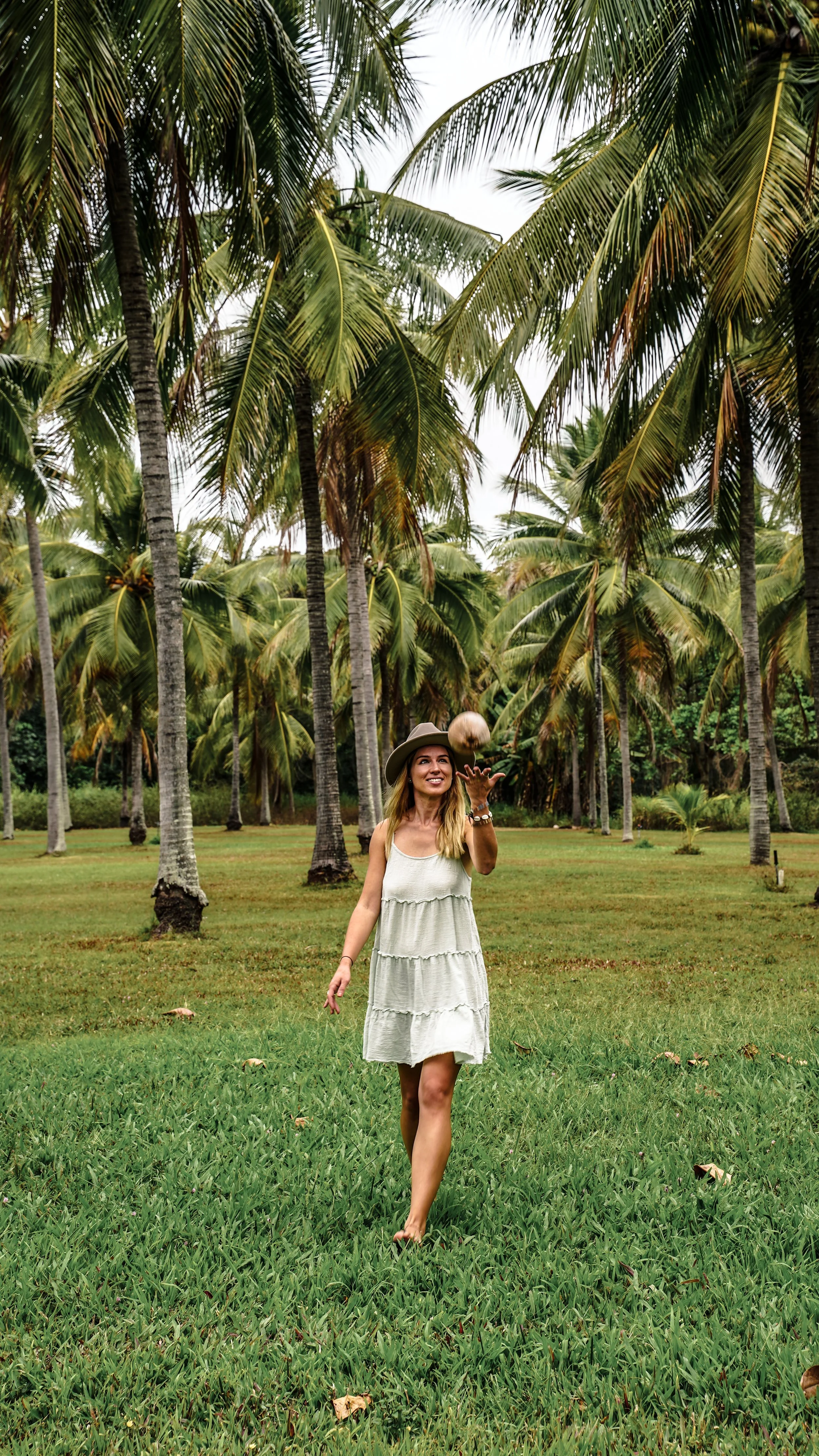 A woman in a white dress and wide-brimmed hat walking barefoot on a grassy field with palm trees in the background, tossing a coconut in the air.