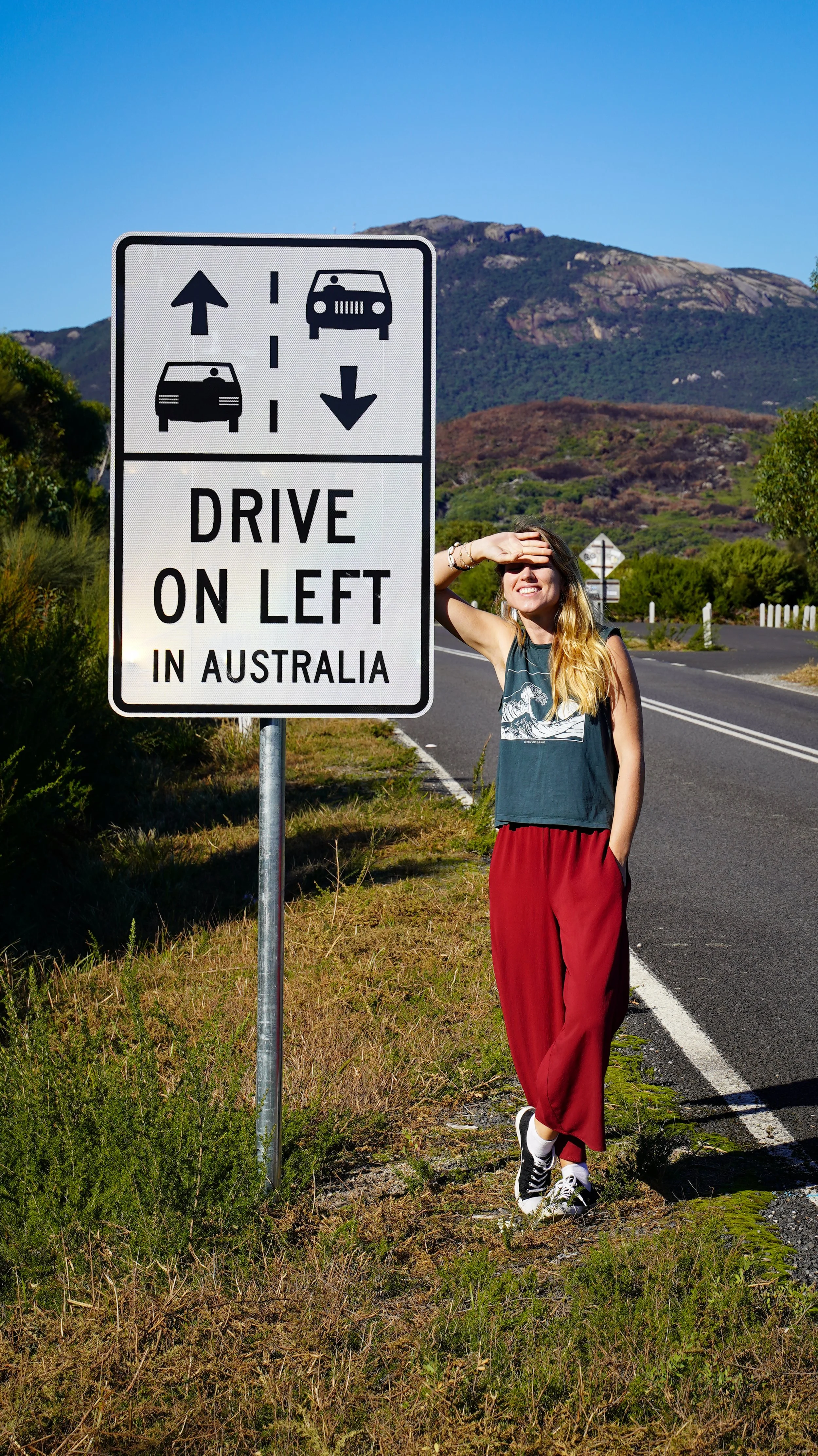 Girl standing next to a road sign that says 'Drive on Left in Australia', shielding her eyes from the sun.