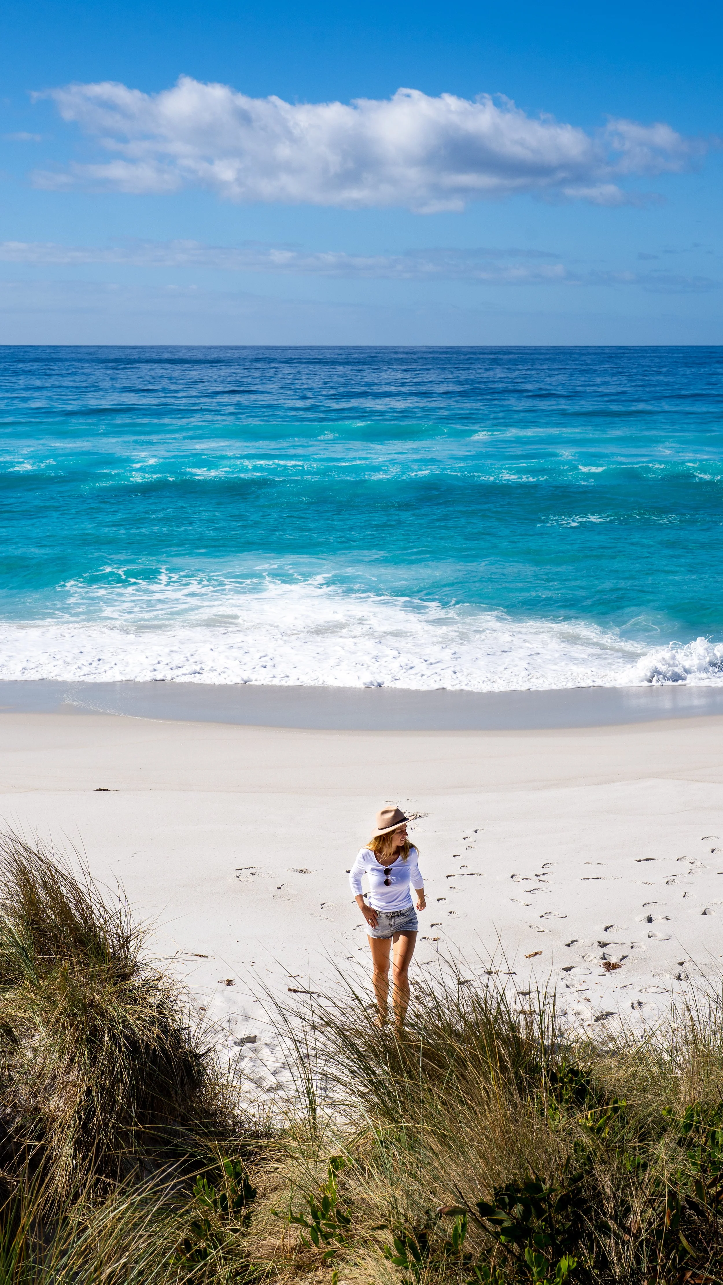 A woman with blonde hair, wearing a beige sun hat, white top, and denim shorts, walking through grassy dunes on a beach towards the ocean with clear blue water and sky with clouds.